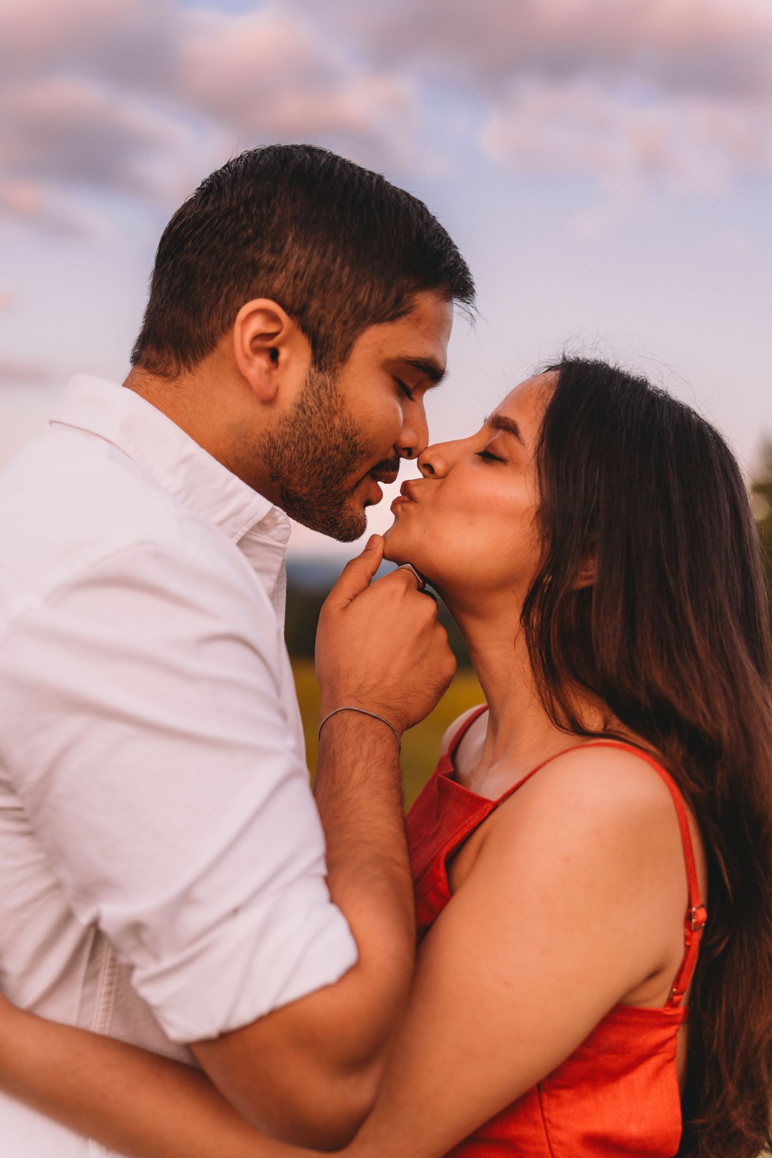 Romantic couple sharing a tender kiss outdoors at sunset with cloudy sky background.