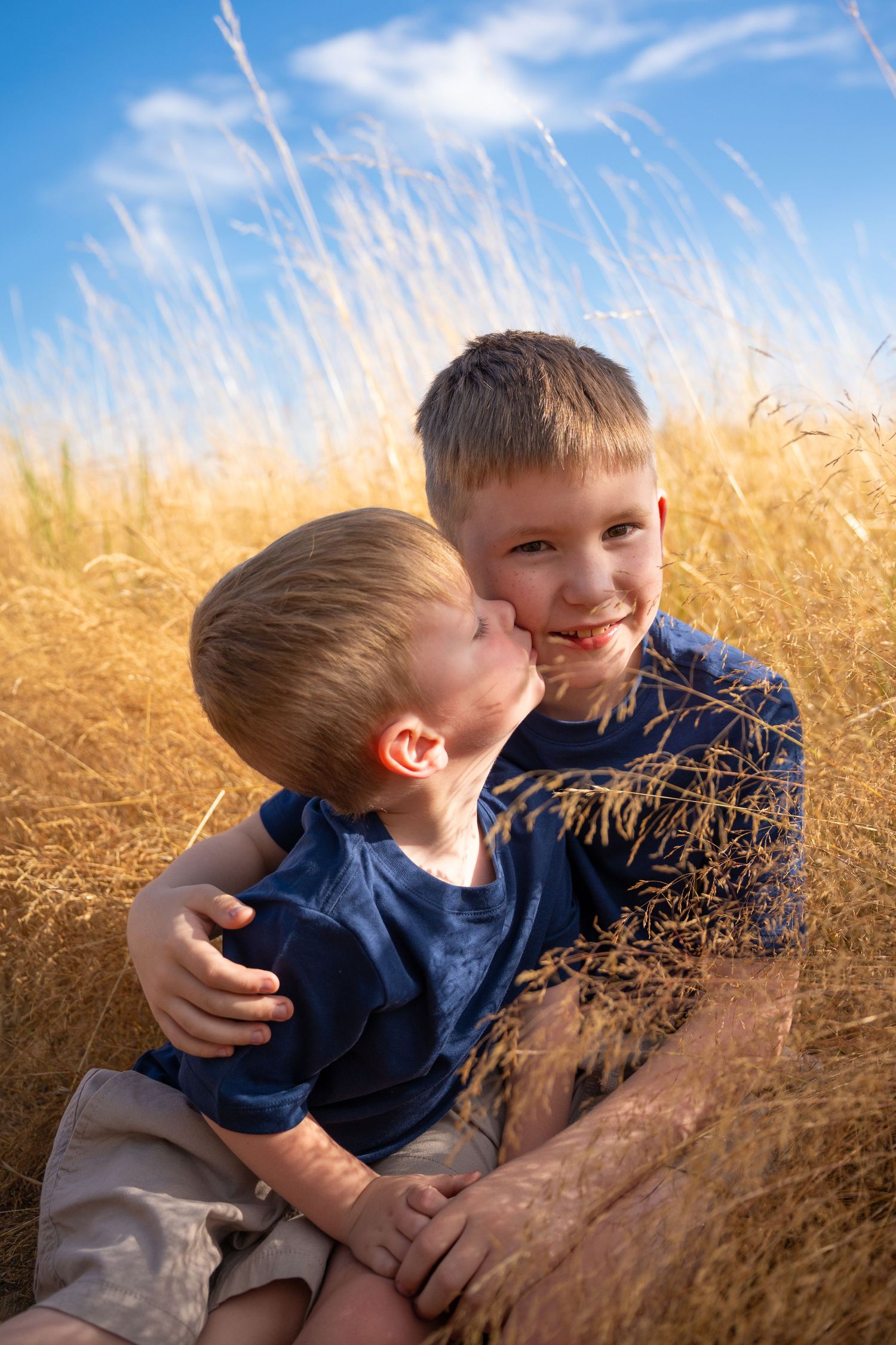 Two young brothers share a sweet kiss in a golden wheat field under a bright blue sky.