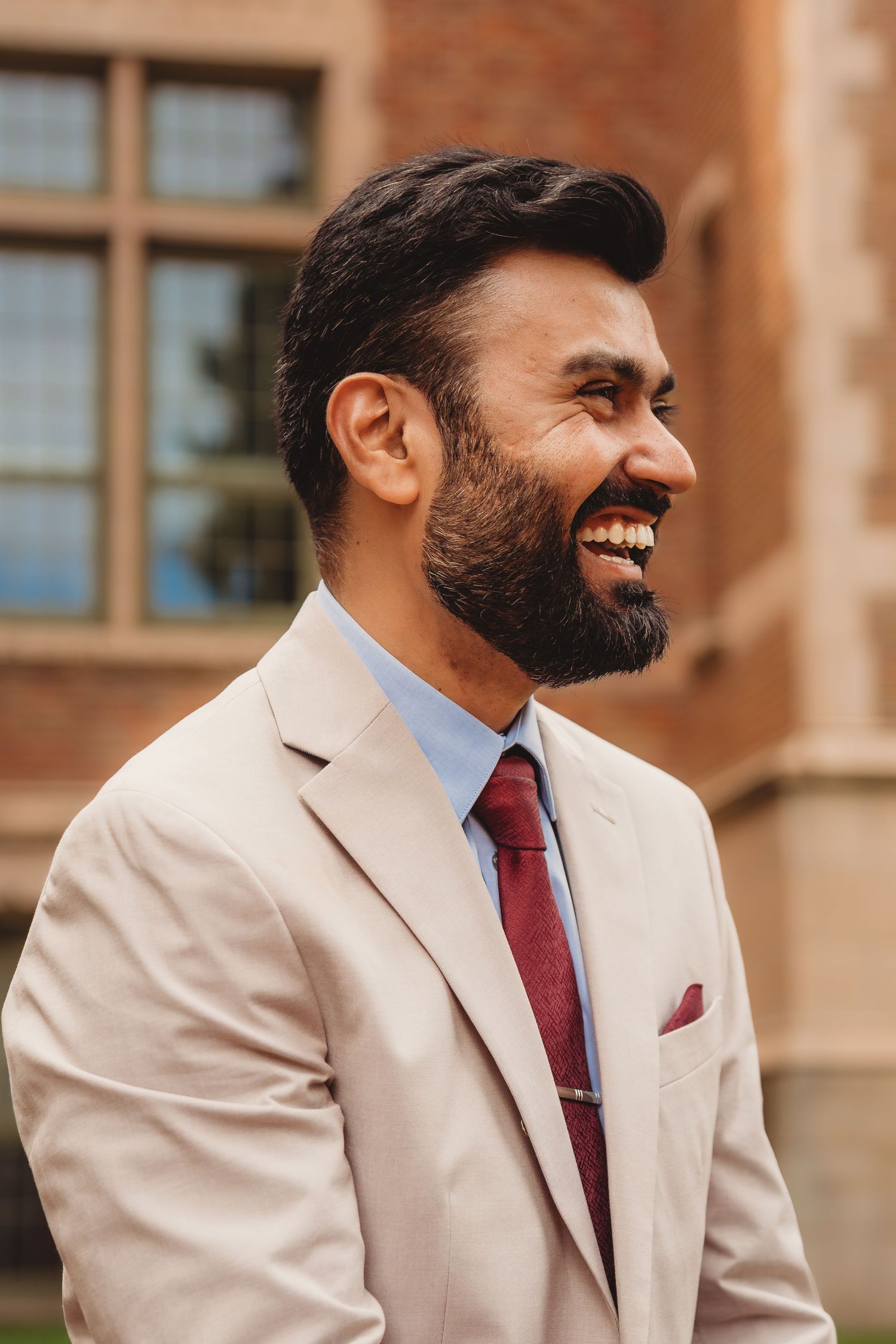 Smiling bearded man in beige suit with burgundy tie and pocket square laughing outdoors near brick building.