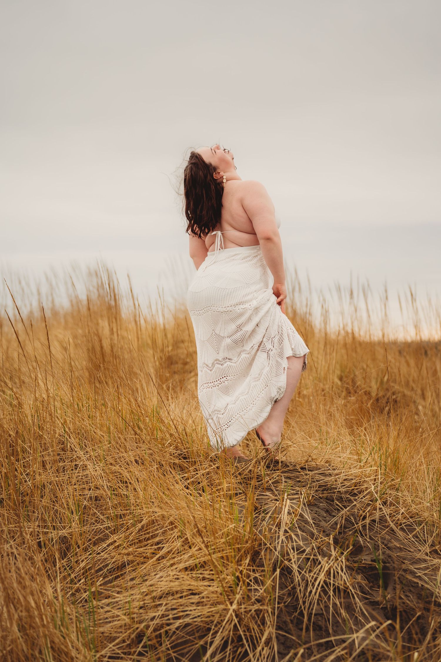 Woman in white lace dress standing in golden grass field, hair blowing in wind, overcast sky backdrop.
