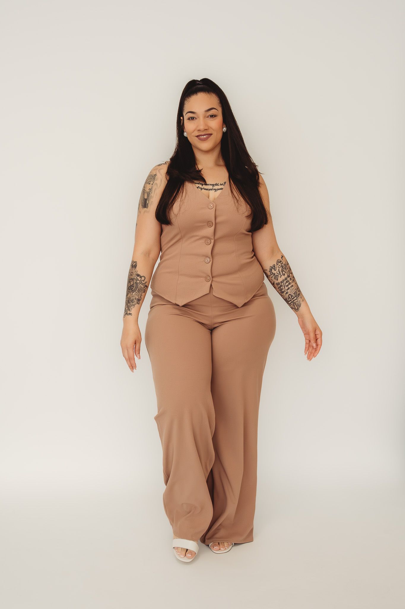 Woman in matching tan vest and wide-leg pants set, showcasing tattoos, posing against white background.