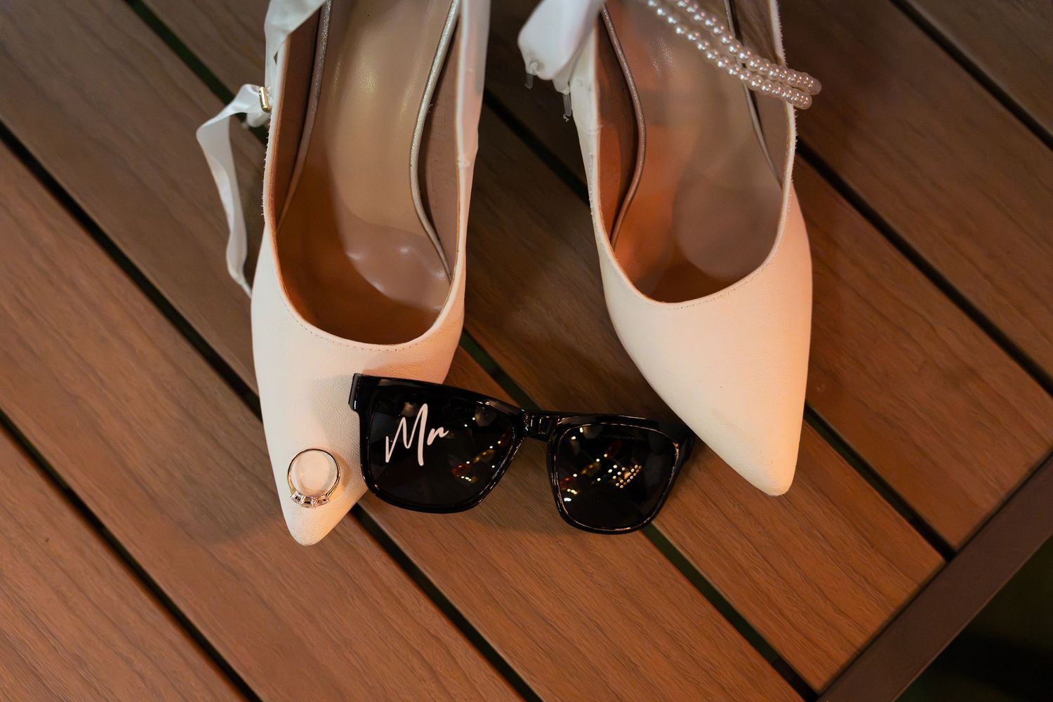 White pointed-toe bridal heels with a pearl strap and a wedding ring, beside black sunglasses marked Mr on a wooden deck.