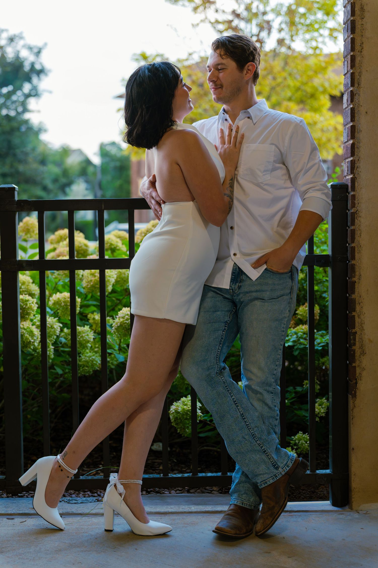 Couple embracing on a balcony by a black railing, she in a white dress and heels, he in a white shirt and jeans, with yellow flowers behind.