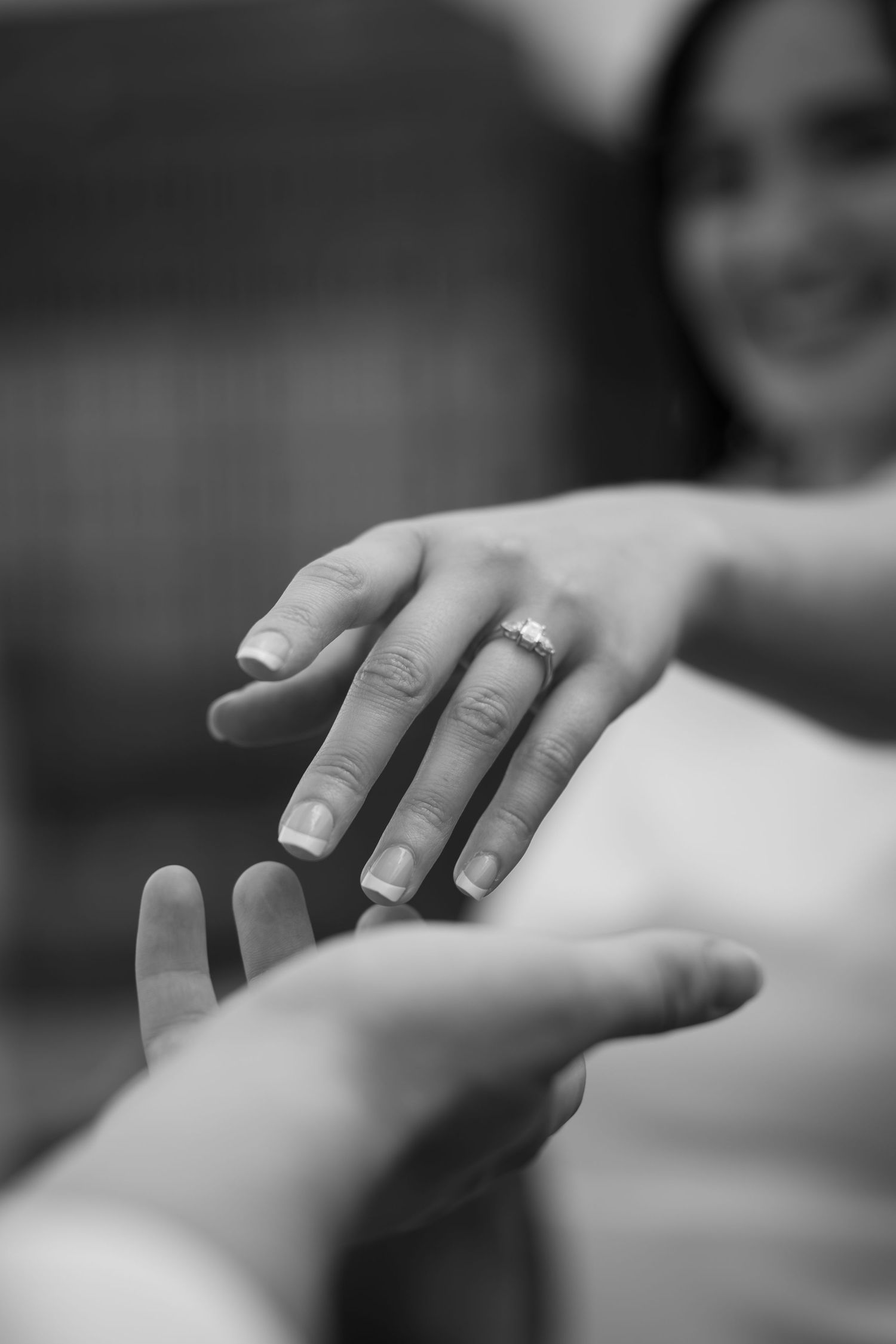 Black and white photo of a smiling woman extending her hand with a diamond engagement ring toward a partner's reaching hand.