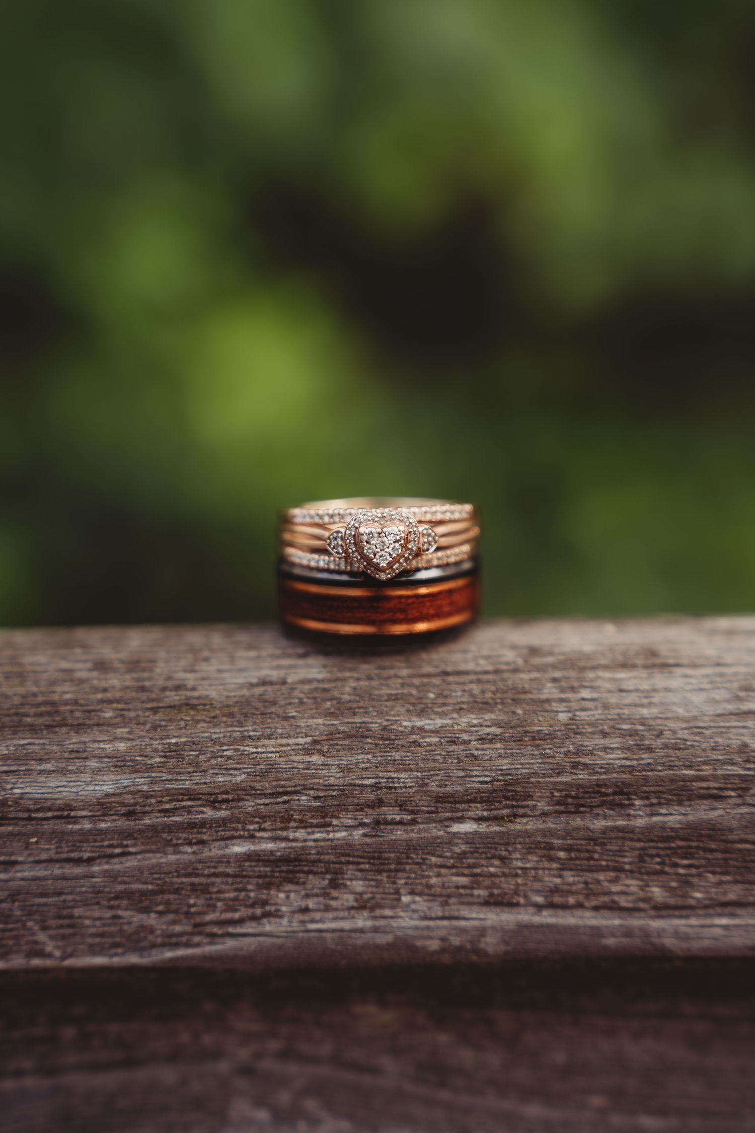 Gold heart diamond engagement ring stacked with wedding bands on rustic wood surface with green bokeh background.