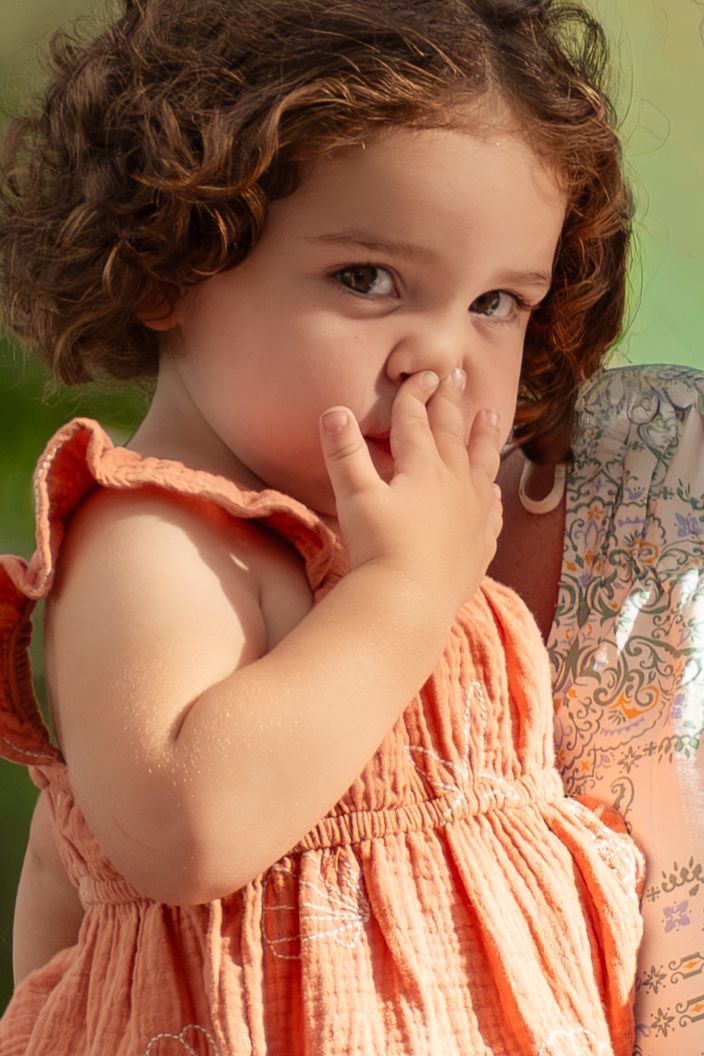 A curly-haired toddler girl in an orange dress covers her mouth with her hand outdoors.