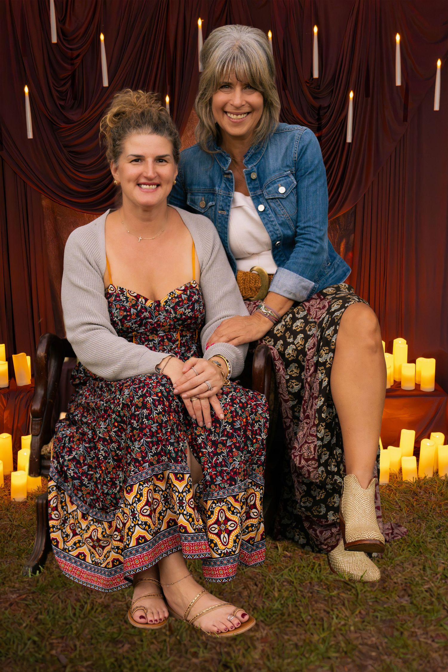 Two smiling women in floral boho dresses pose together outdoors on grass in front of a red curtain and glowing candles.