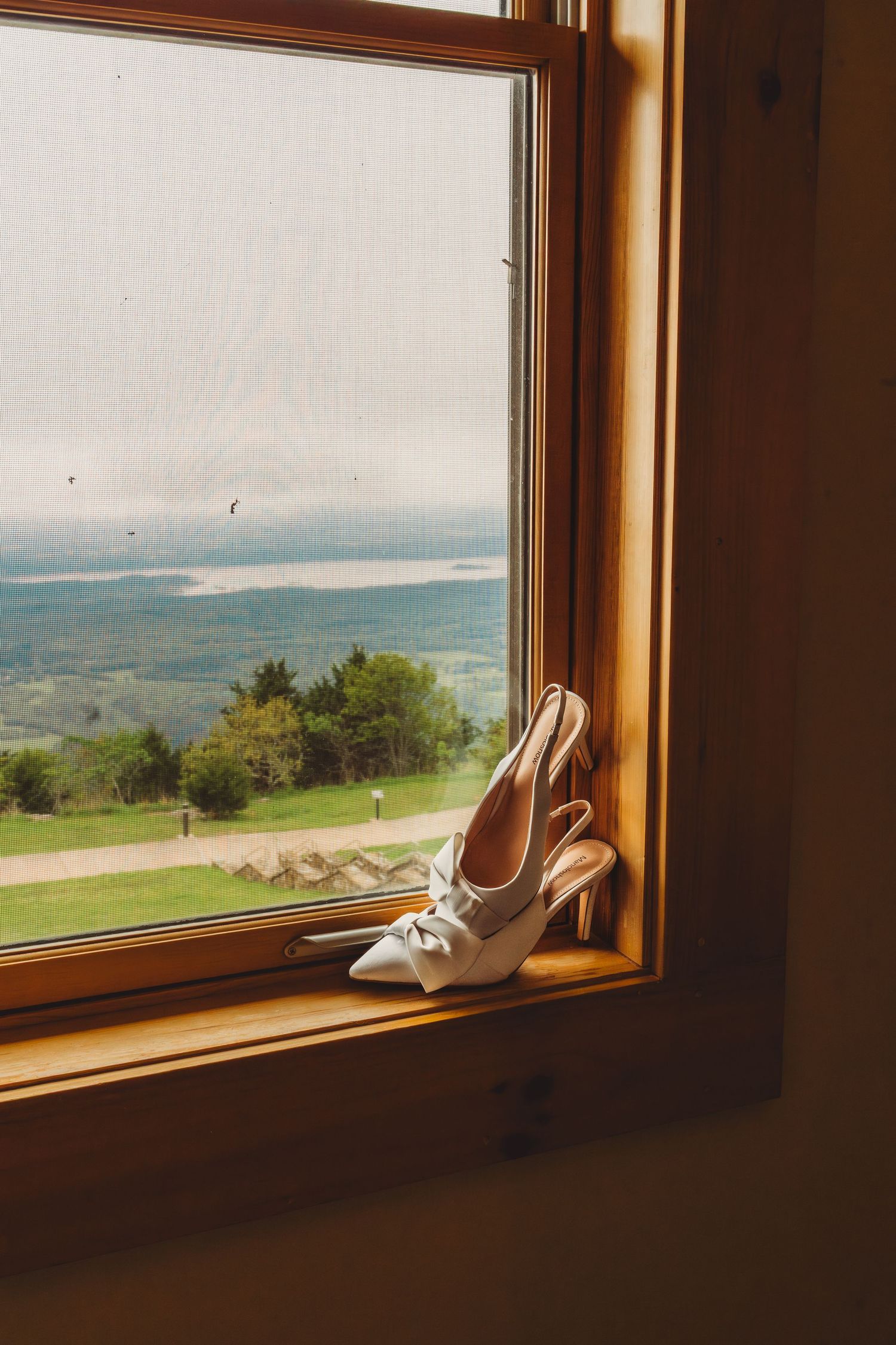 White bridal heels with bow detail resting on a wooden windowsill overlooking a scenic green landscape. Arkansas