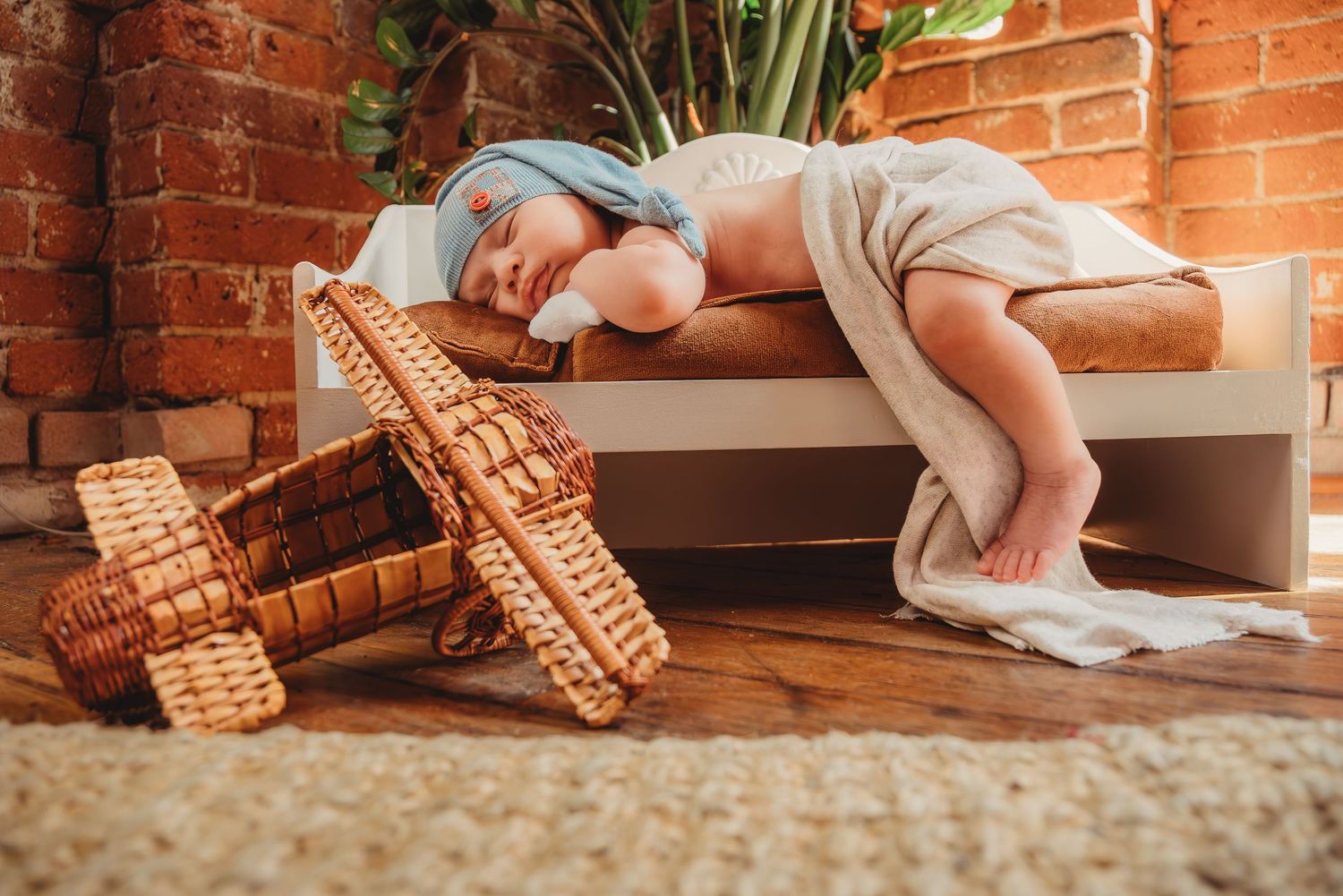 Sleeping newborn baby wrapped in soft blanket rests on bench beside wicker toy airplane.