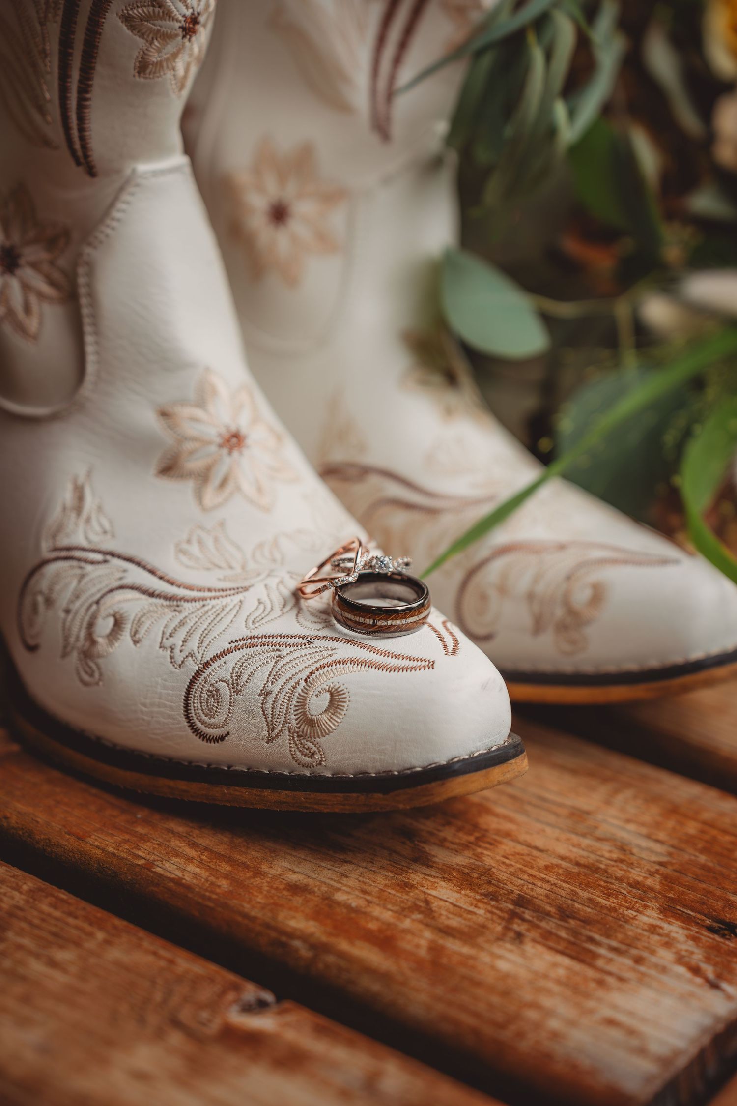 White cowboy boots with floral embroidery display two wedding rings on a rustic wooden surface with greenery background.