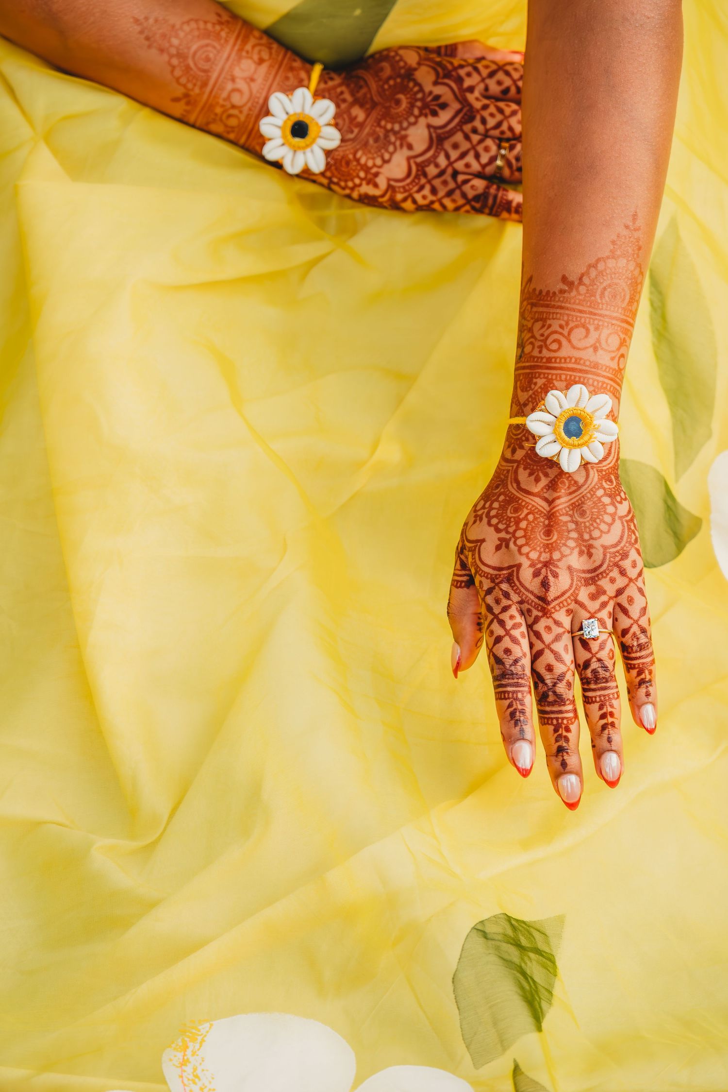 Intricate henna mehndi designs on bride's hands adorned with white daisy bracelets against yellow fabric.