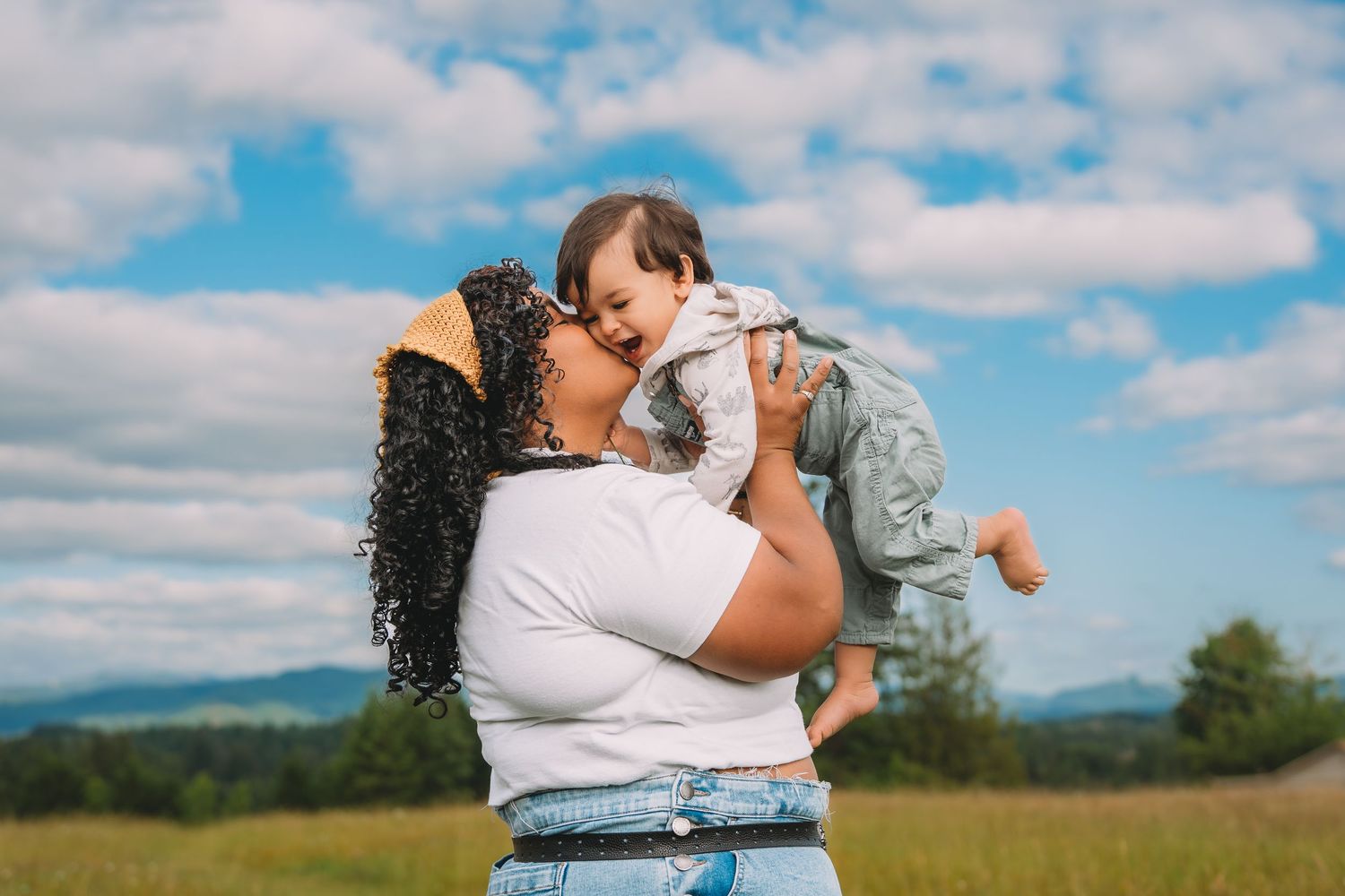 A mother joyfully lifts her baby outdoors in a green field under a blue sky with clouds.