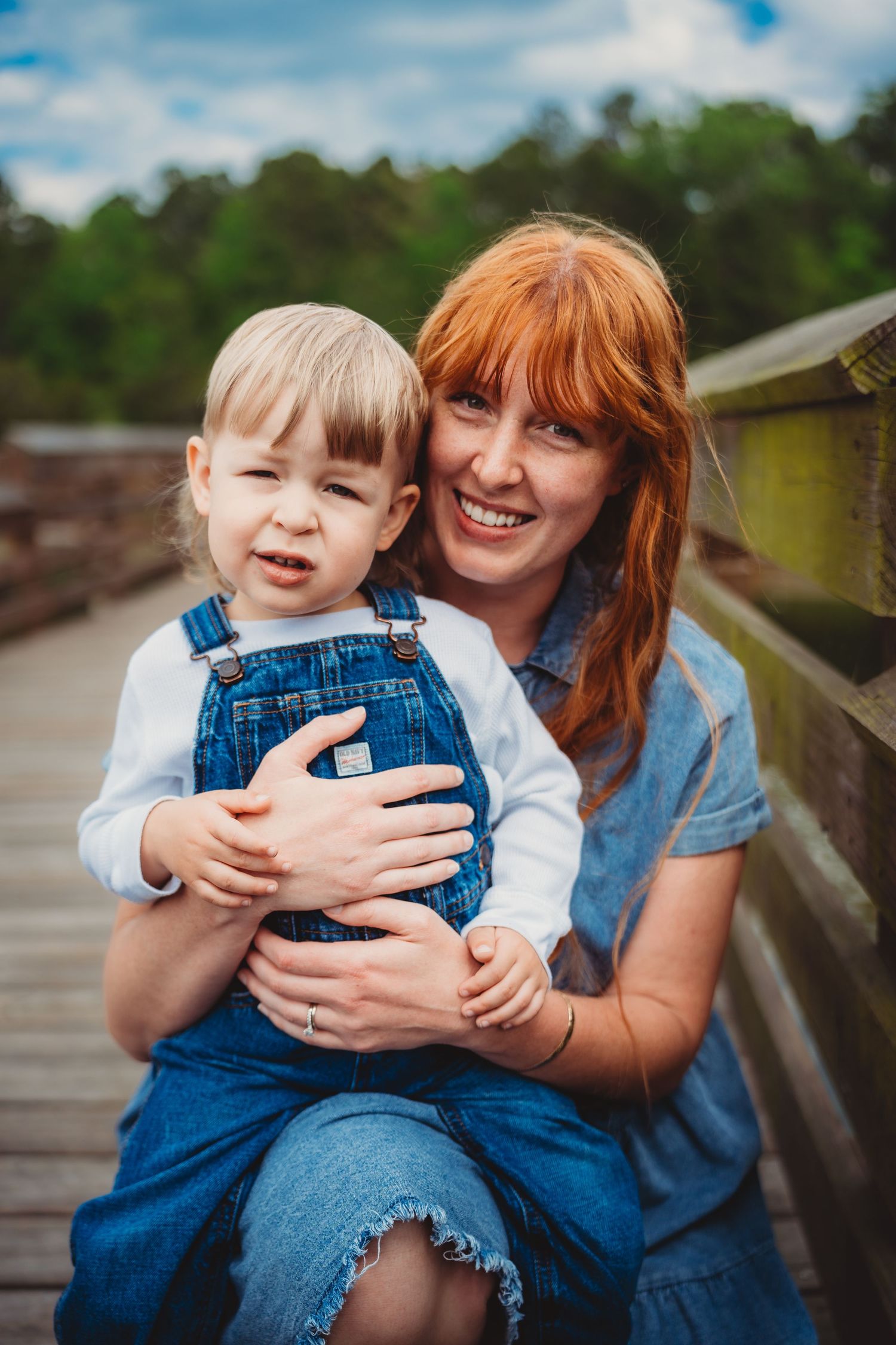 A smiling redhead woman hugs a toddler in denim overalls on a wooden bridge surrounded by green trees.