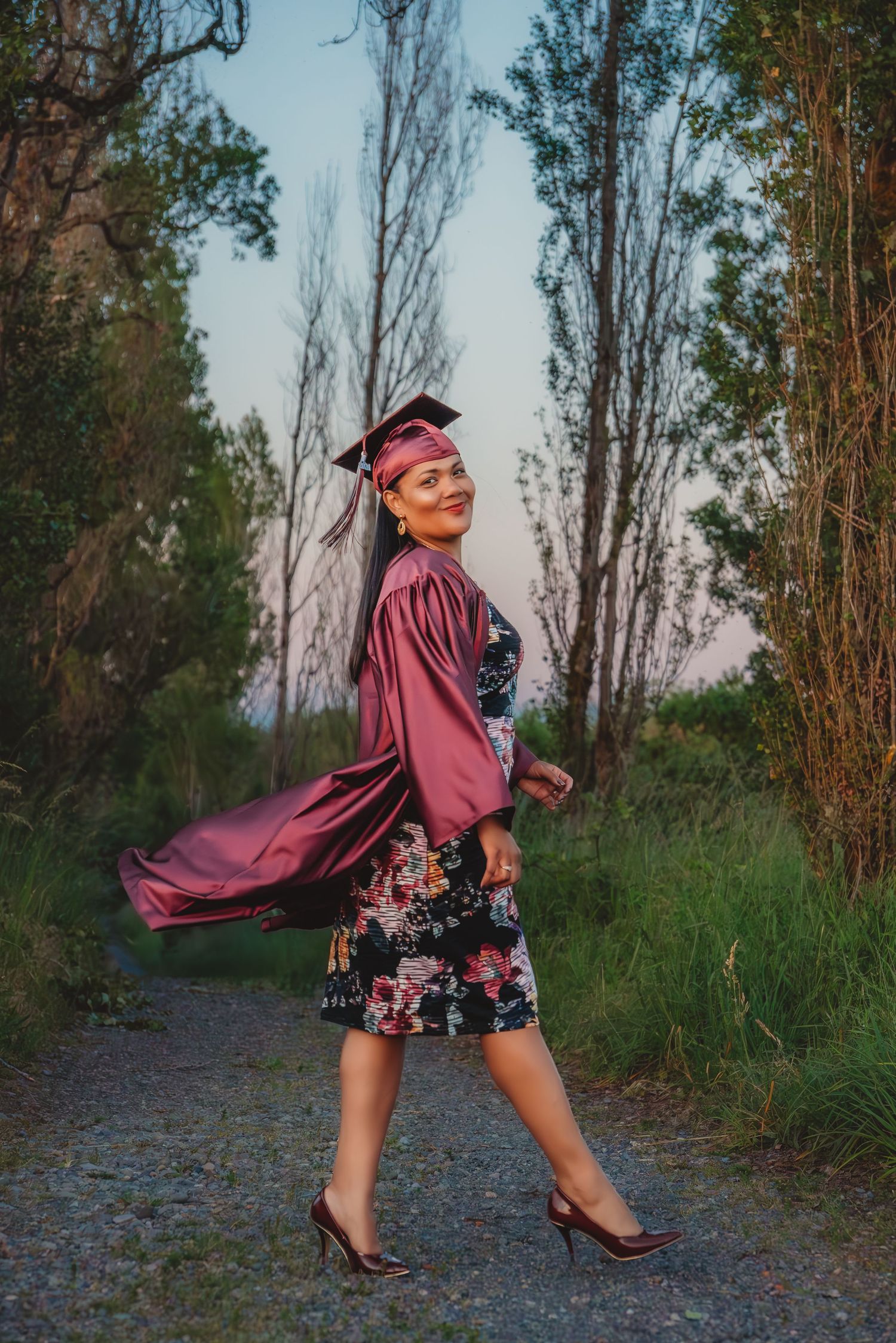 Smiling graduate in burgundy cap and gown walking on tree-lined path at sunset. South Carolina