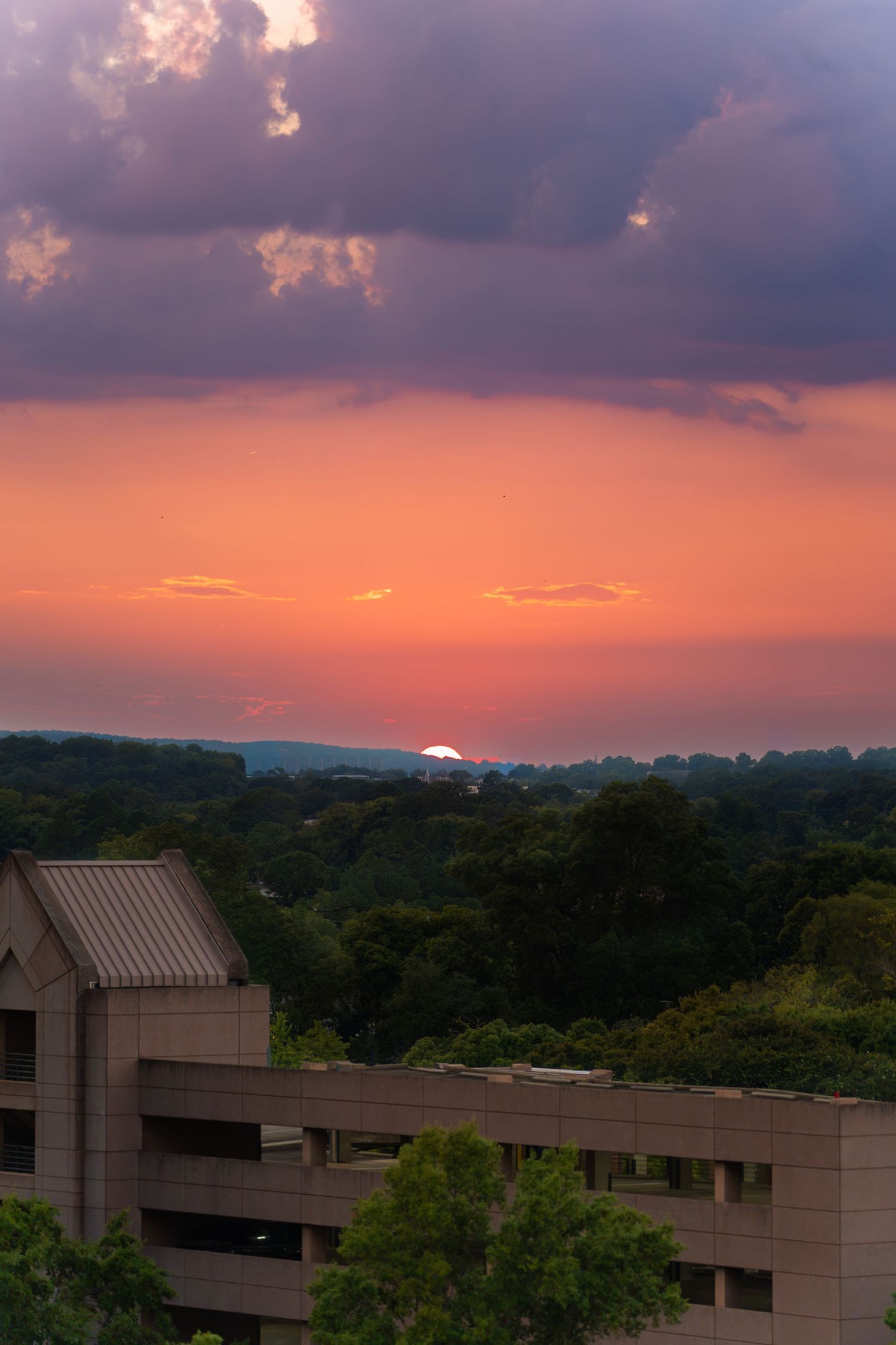 Vibrant orange and purple sunset sky with dramatic clouds over a lush green tree canopy and a modern tan building in the foreground.