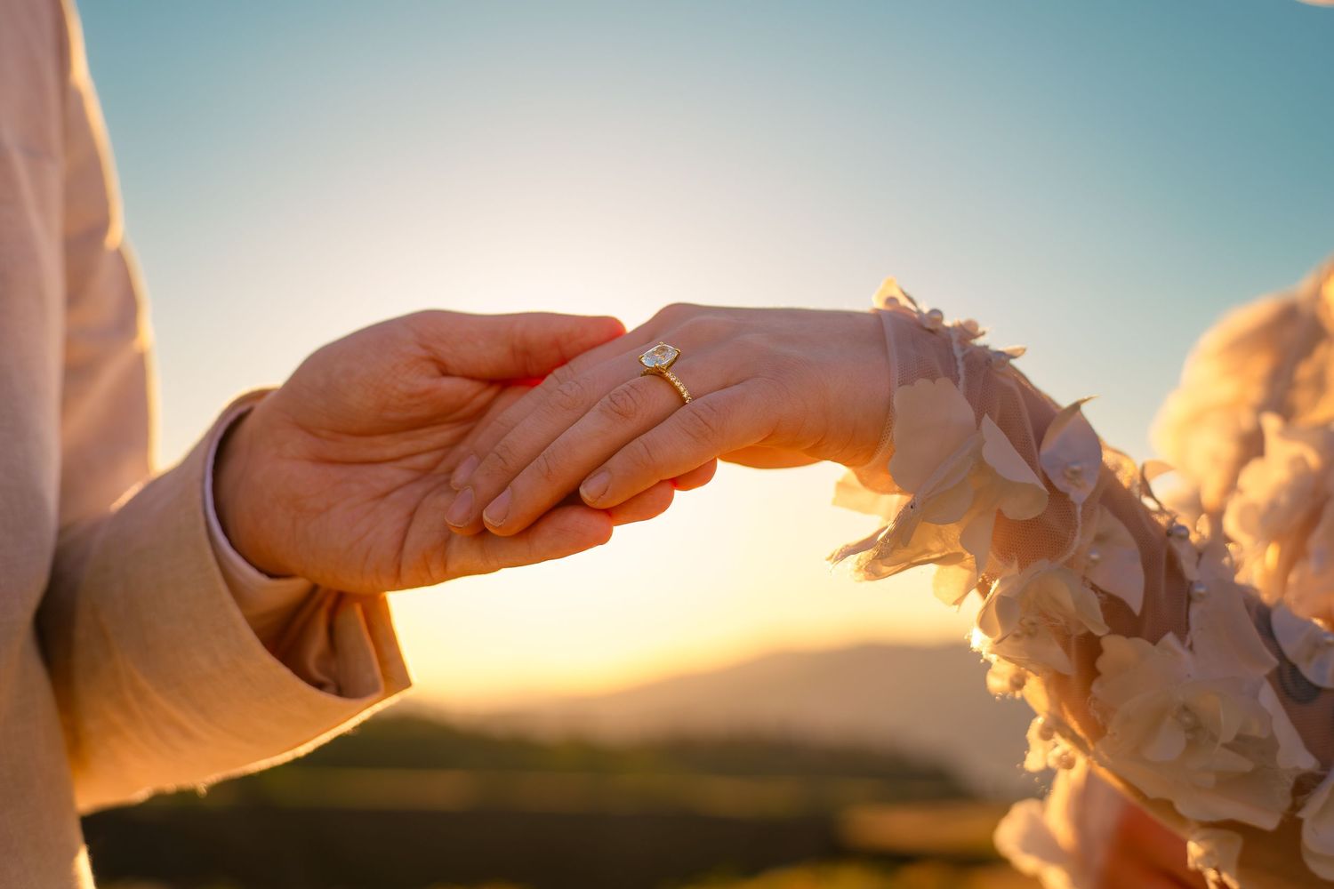 Close-up of couple holding hands at sunset with wedding ring visible against scenic mountain backdrop.