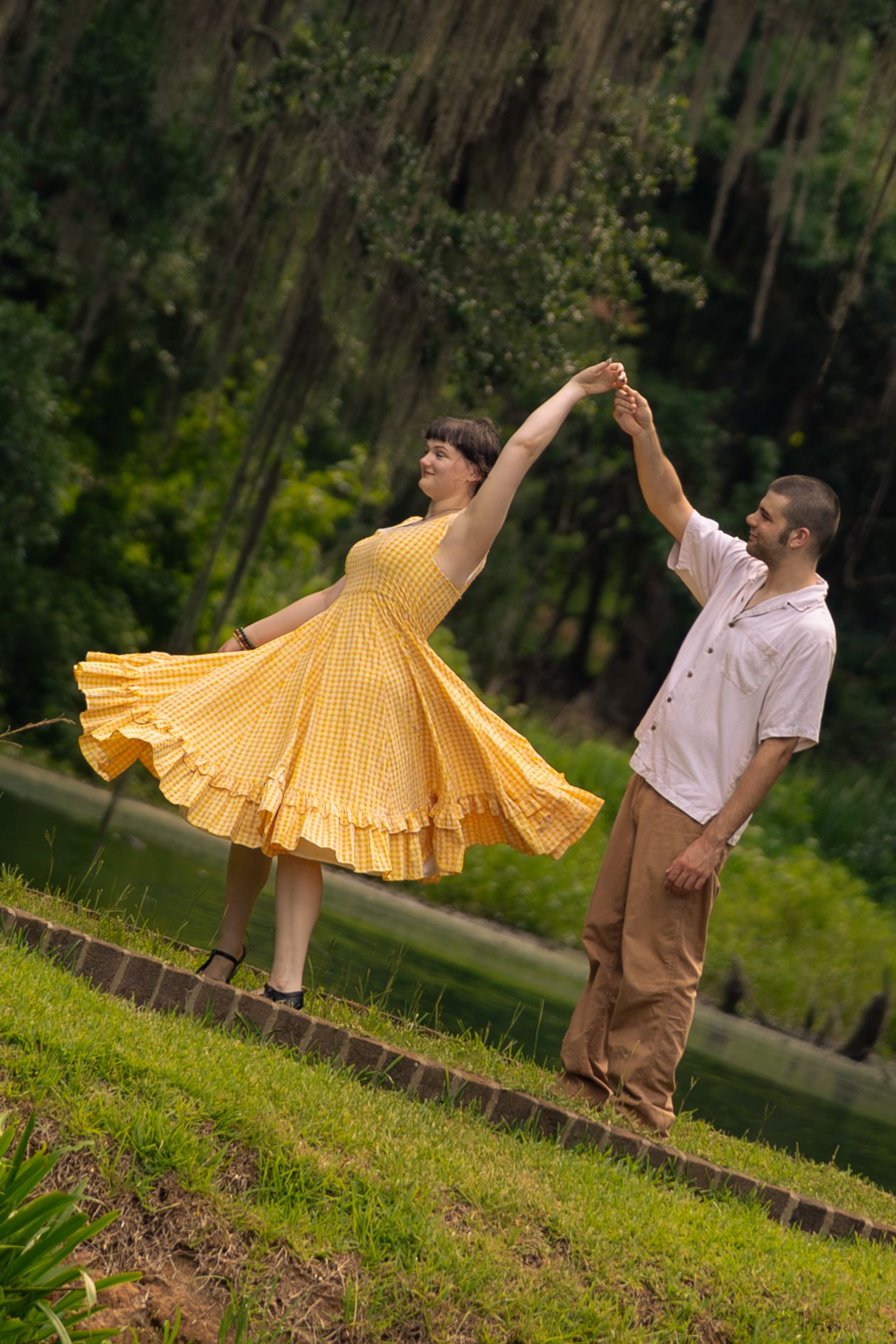 A couple dances outdoors, woman in flowing yellow dress twirling with man in white shirt near a pond.