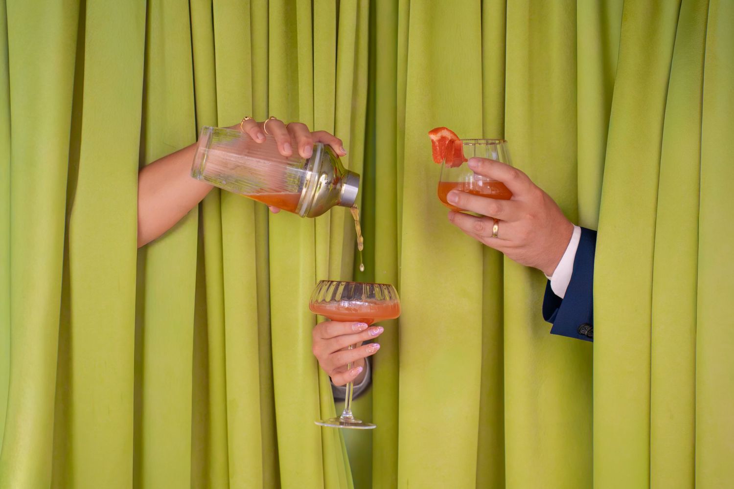 Two hands emerging from green curtains, one pouring a cocktail shaker into a glass, the other holding a garnished drink.