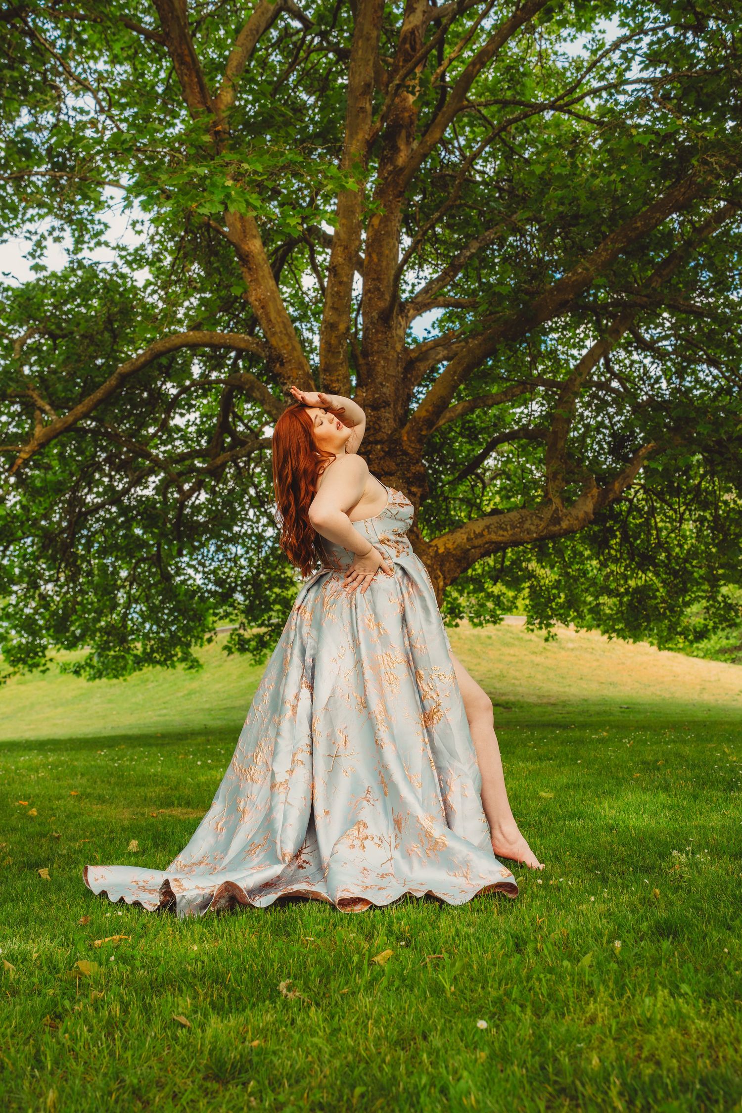 A redhead woman in a floral ball gown poses gracefully barefoot on green grass beneath a large tree.