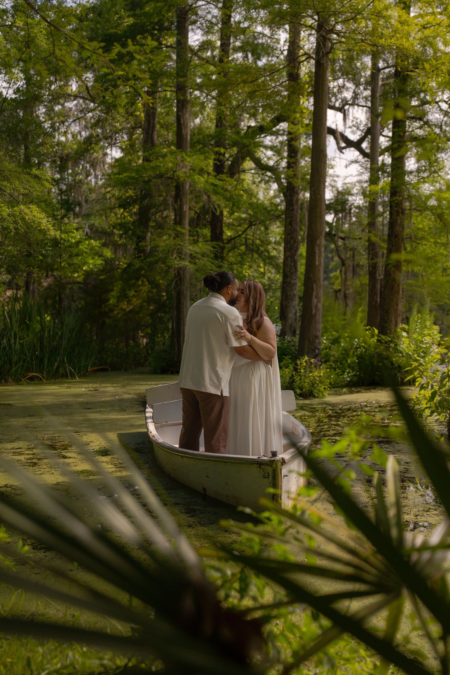 Couple sharing a romantic kiss in a white canoe on a serene, moss-covered swamp surrounded by lush green trees.