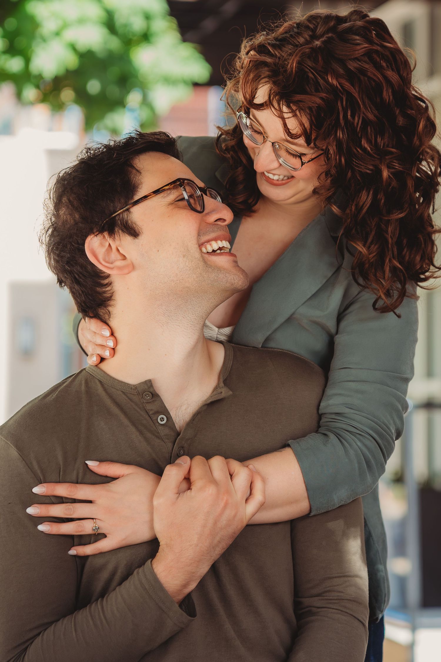 A smiling couple with glasses sharing a warm embrace outdoors, the woman hugging the man from behind as they laugh together.