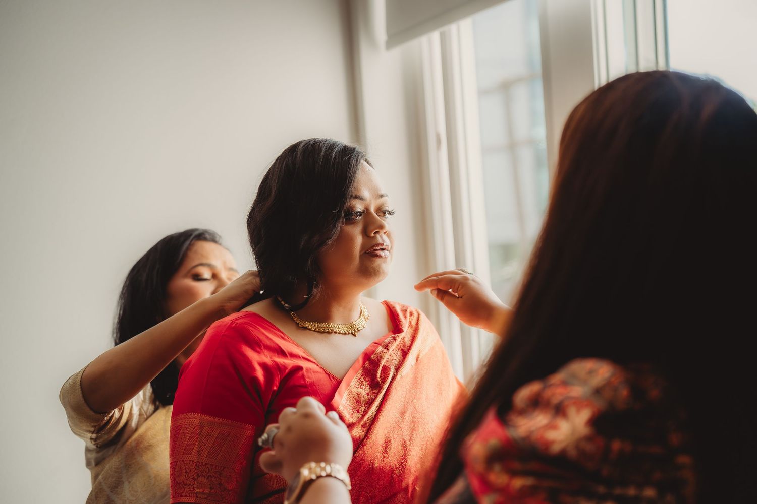 Woman in red saree being helped by two others near a bright window during a traditional Indian wedding preparation.