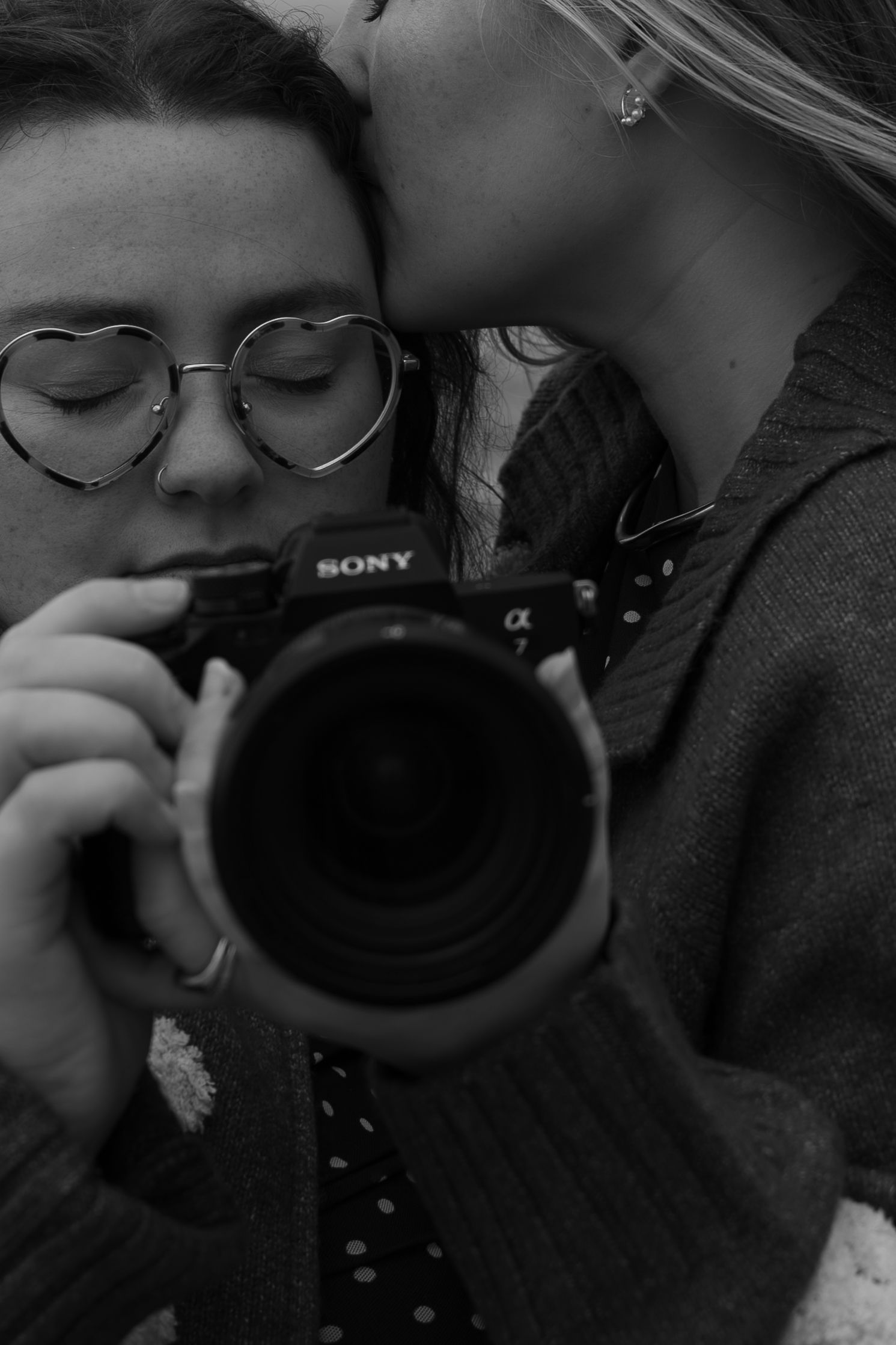Black and white photo of two women, one kissing the other while she looks through a Sony alpha camera.