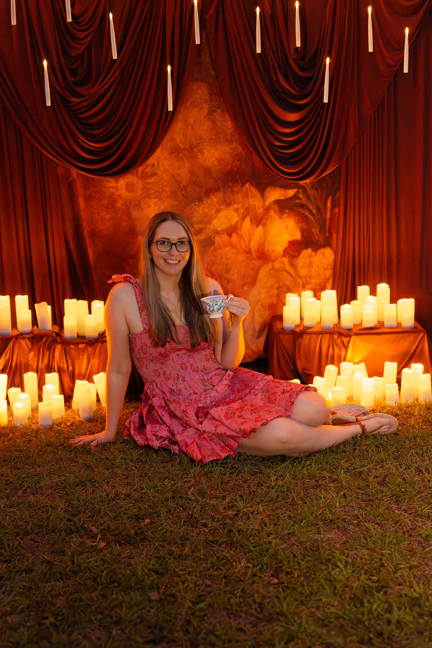 Woman in glasses and a pink floral dress sits on grass holding a teacup, surrounded by glowing candles and red draped curtains.