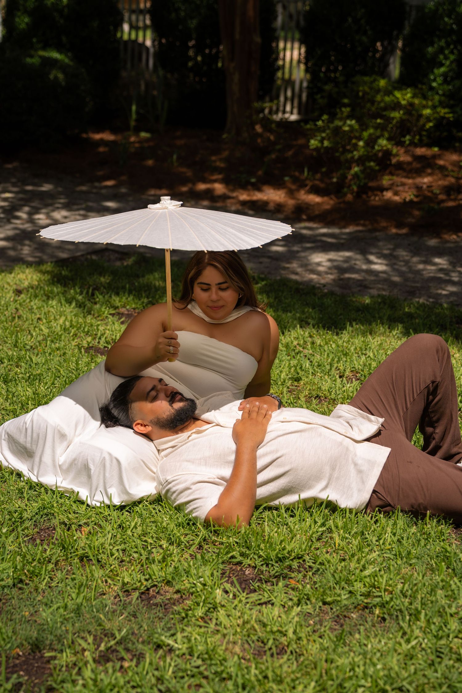Couple relaxing on grass in park, woman holding white parasol umbrella over man lying on her lap.