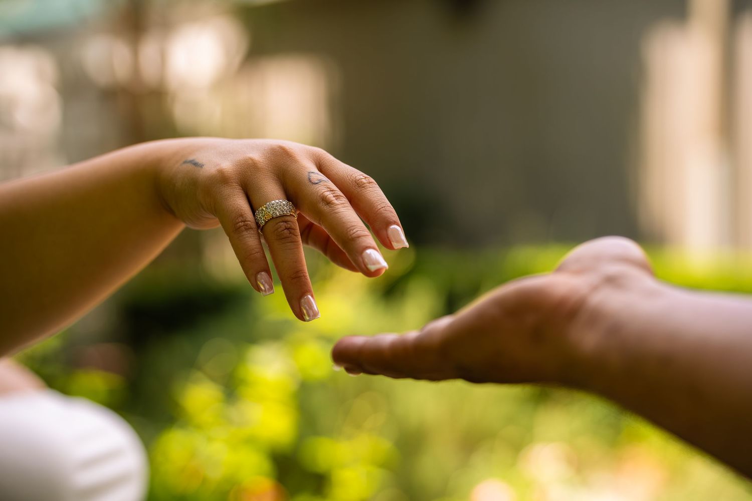 Two hands reaching toward each other, one wearing a gold ring, with lush green bokeh background.