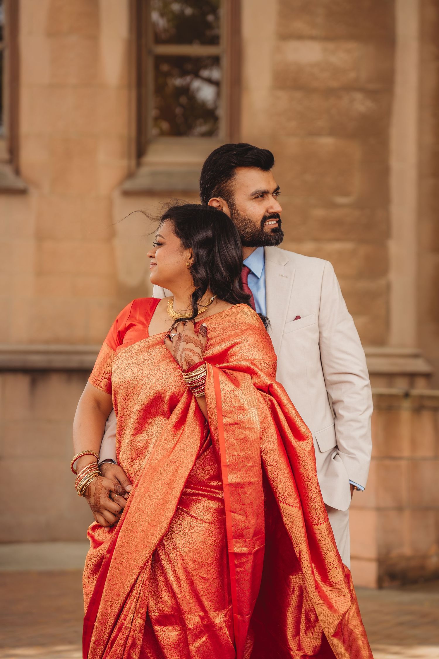 Couple in traditional Indian wedding attire, woman in orange saree and man in white suit, posing elegantly outdoors.