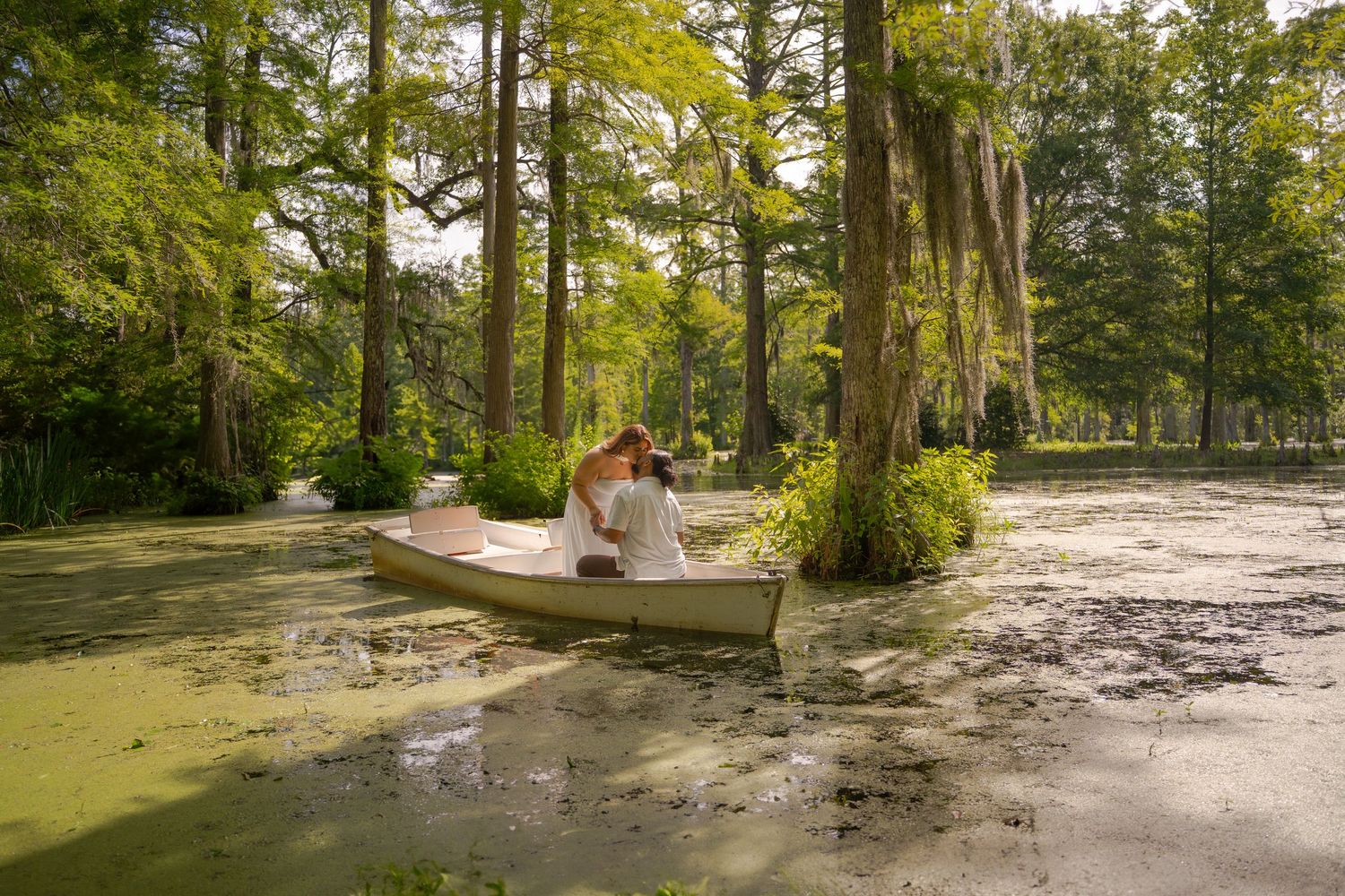Couple embracing in a white canoe surrounded by cypress trees and green algae in a serene swamp setting.