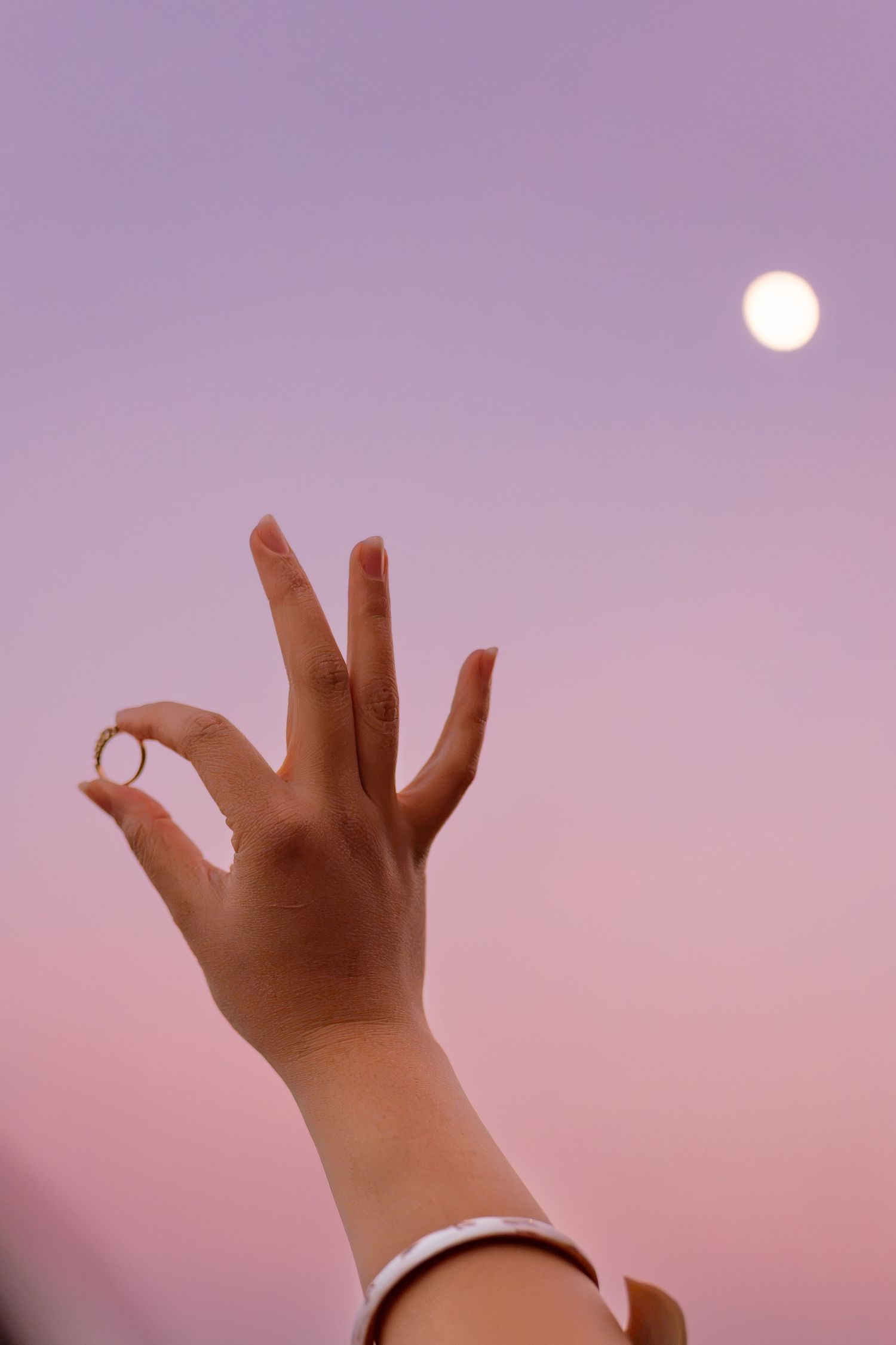 A hand with a gold ring raised against a pink and purple twilight sky with a full moon in the background.