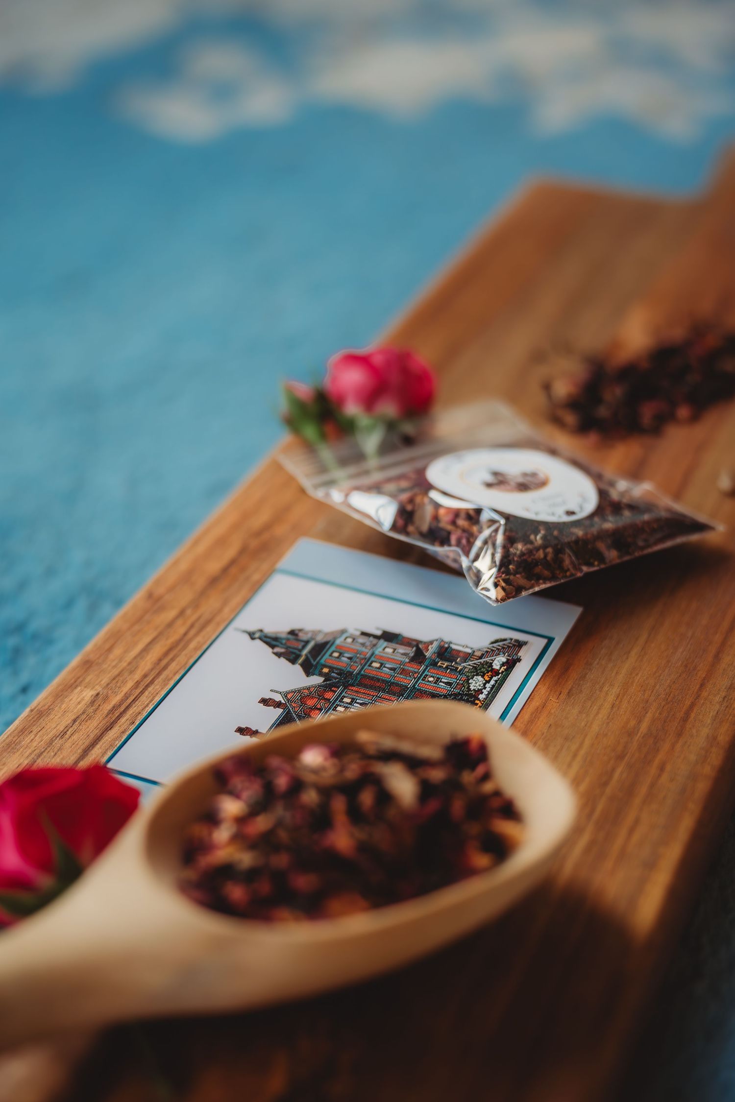 Wooden spoon with dried hibiscus tea leaves beside a postcard and small bag of tea on a rustic wooden board.