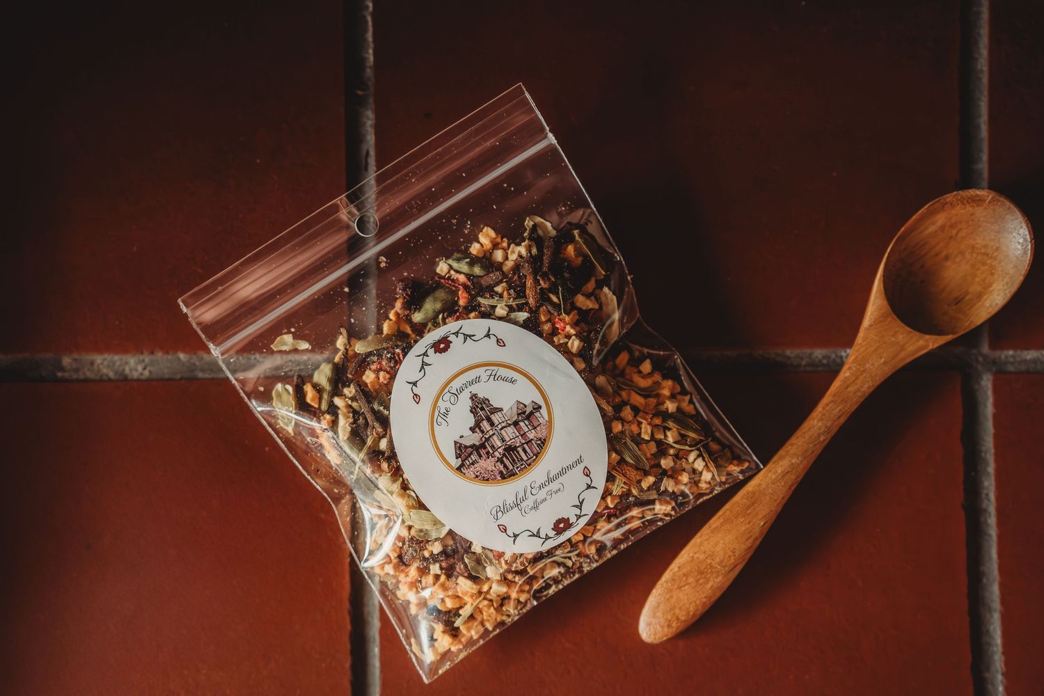 A small ziplock bag of mixed spices with a branded label sits on terracotta tiles beside a wooden spoon.