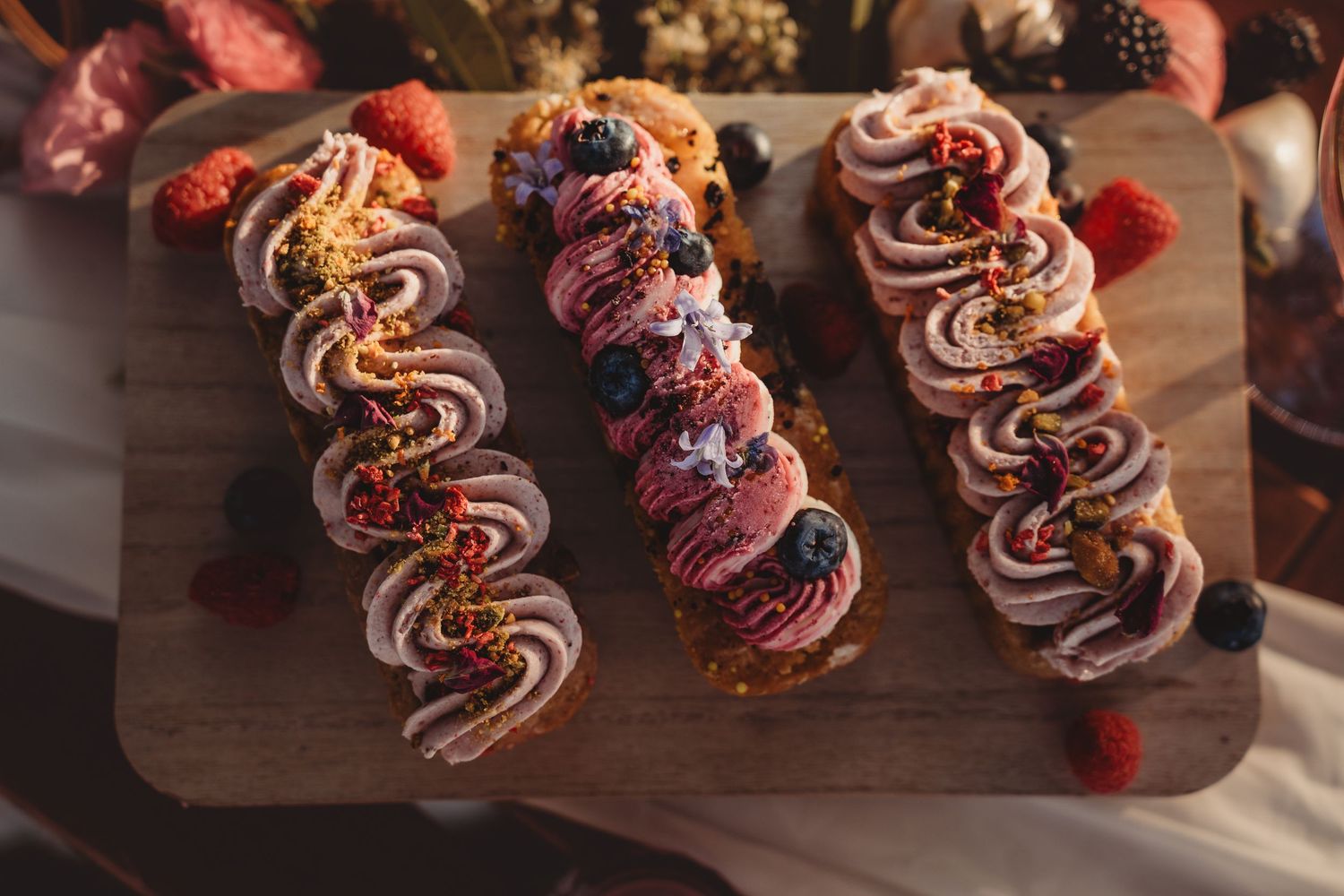 Three elegant berry-topped pastry eclairs with pink frosting on a wooden board, garnished with raspberries and blackberries.