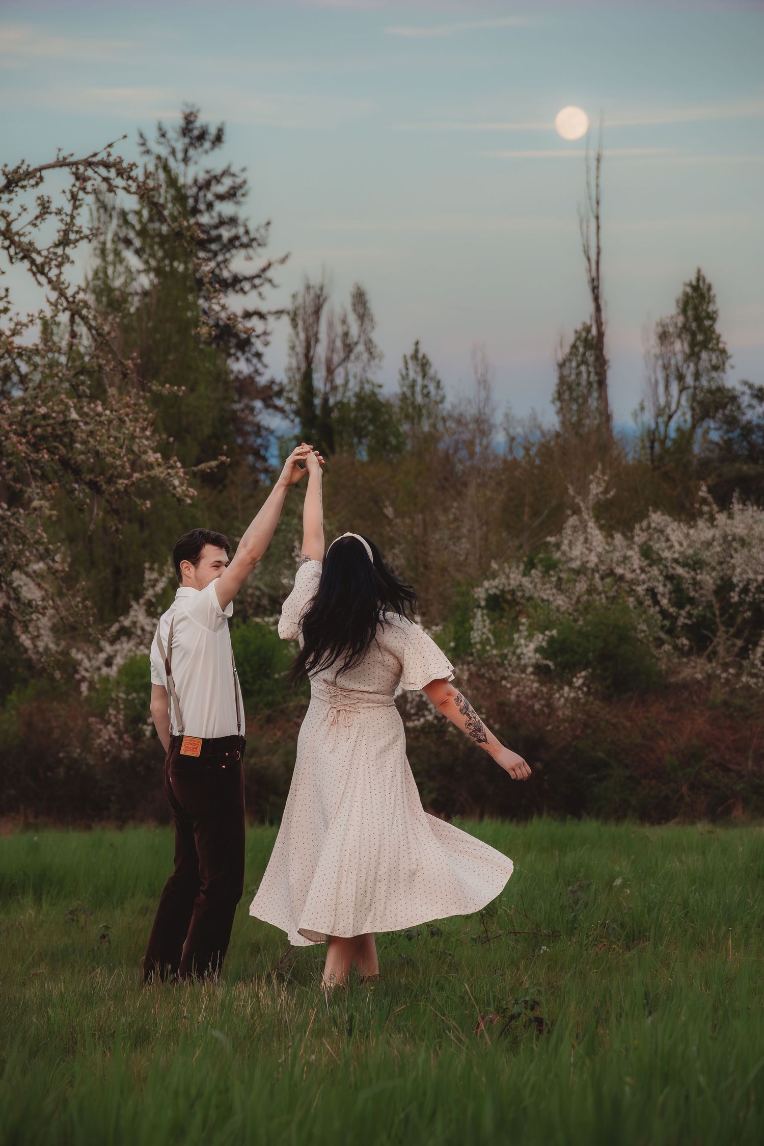 Couple dancing in moonlit field, woman in flowing white dress twirls under full moon at dusk.