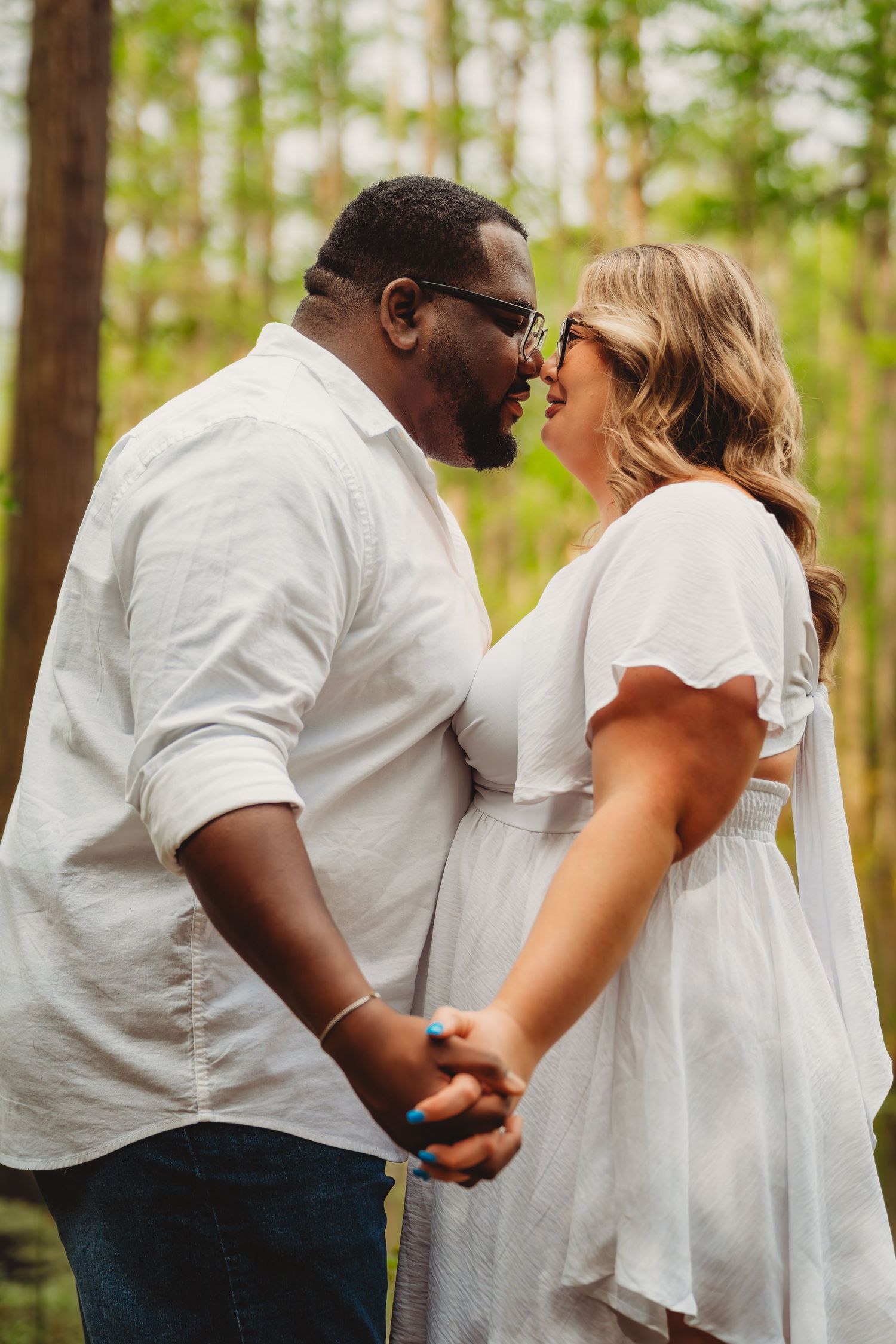 Interracial couple holding hands and smiling at each other in white outfits among tall trees in a forest. cypress gardens