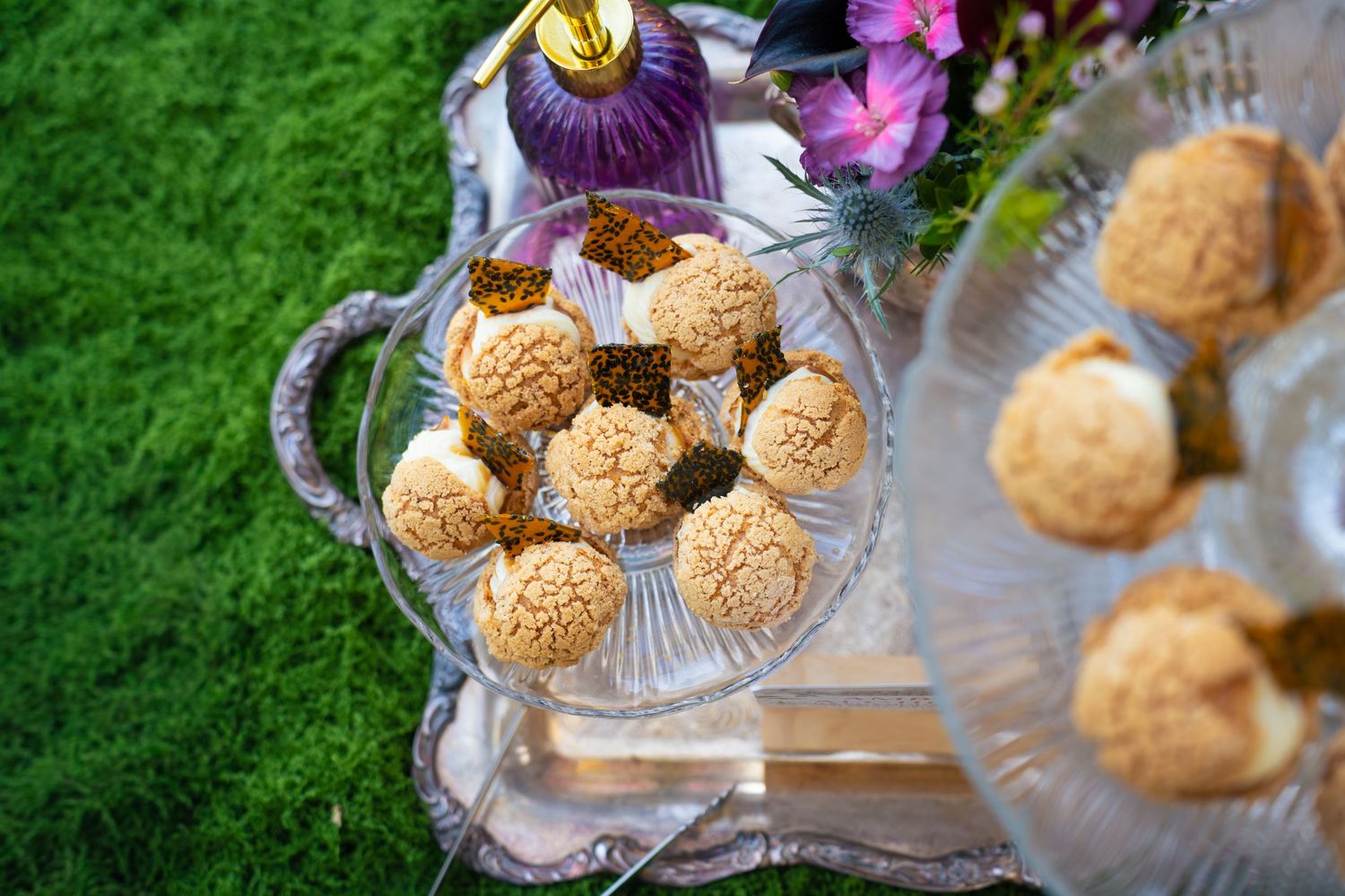 Crunchy craquelin-topped cream puffs on a glass dessert stand with ornate silver tray, purple flowers and a perfume bottle on green grass.