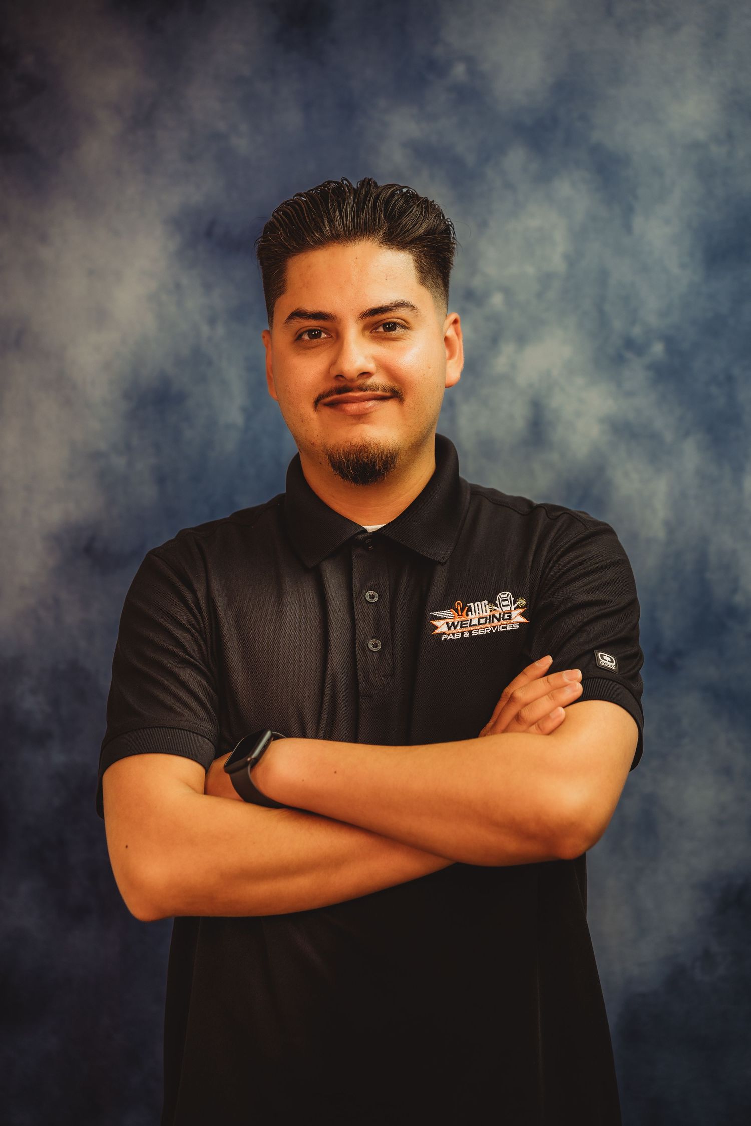 Young man in black polo shirt with arms crossed smiling confidently against a blue studio backdrop.