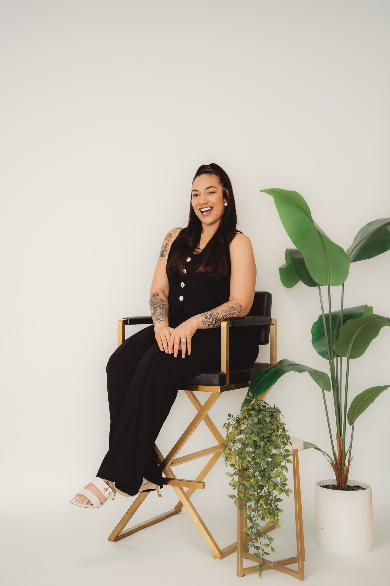 Tattooed woman in black jumpsuit laughing while seated on gold folding chair next to indoor plants.