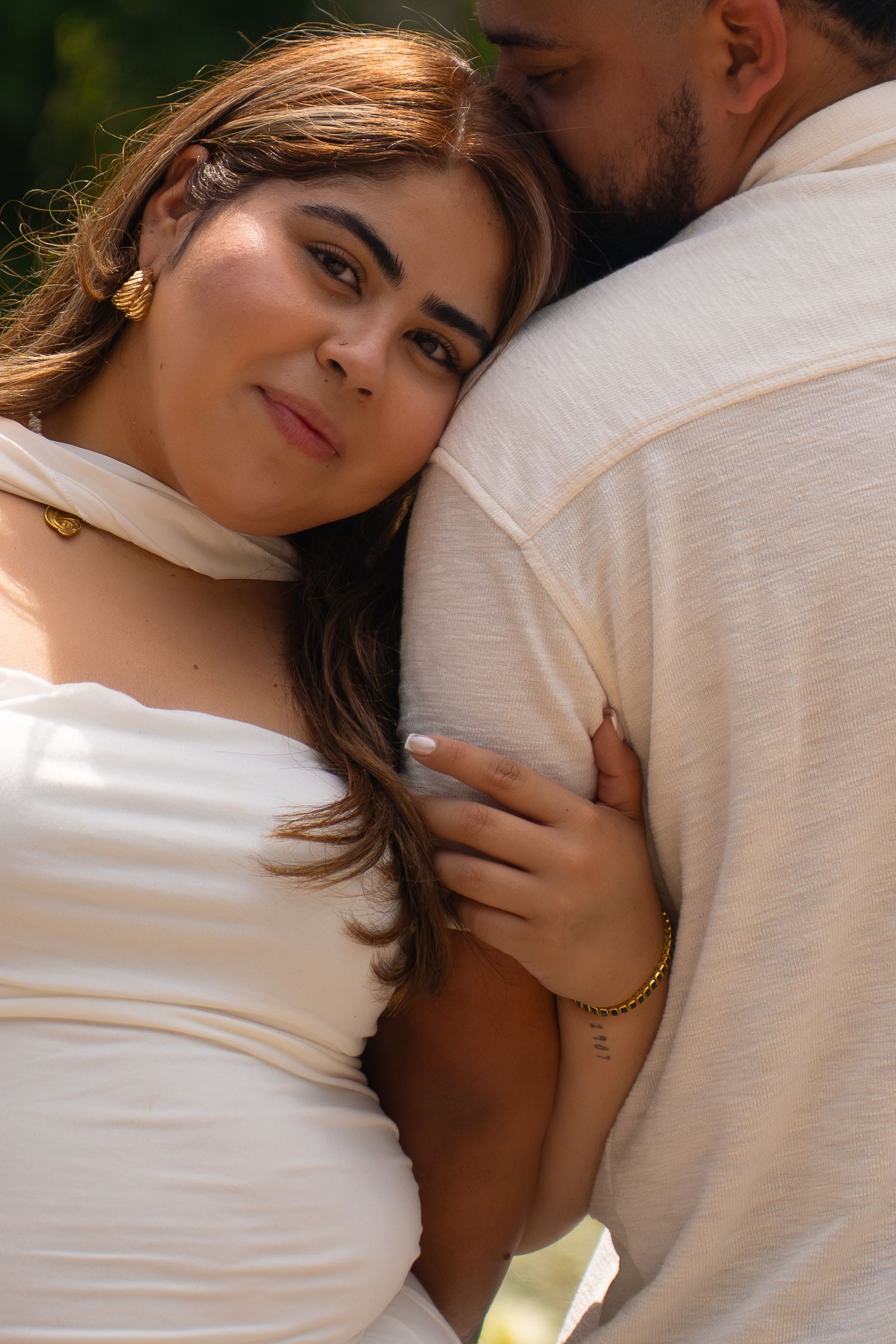 A woman in a white dress rests her head on a man's shoulder, embracing him tenderly outdoors.