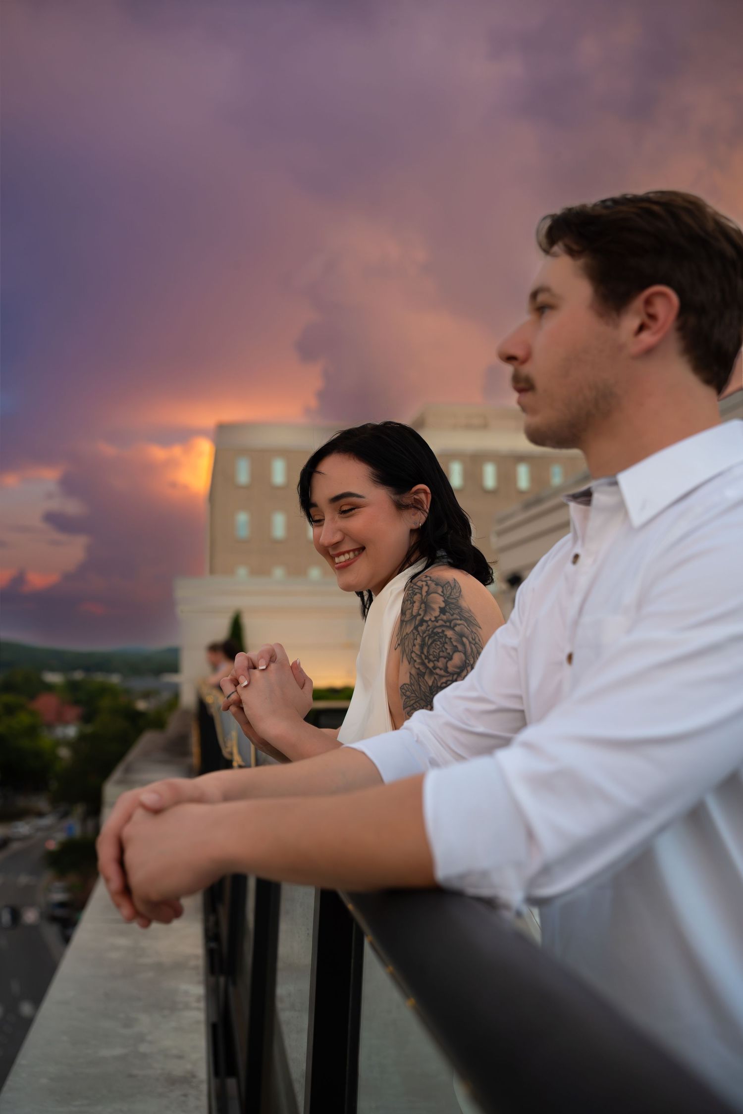 Smiling couple leaning on a rooftop railing overlooking the city beneath a dramatic purple and orange sunset sky.