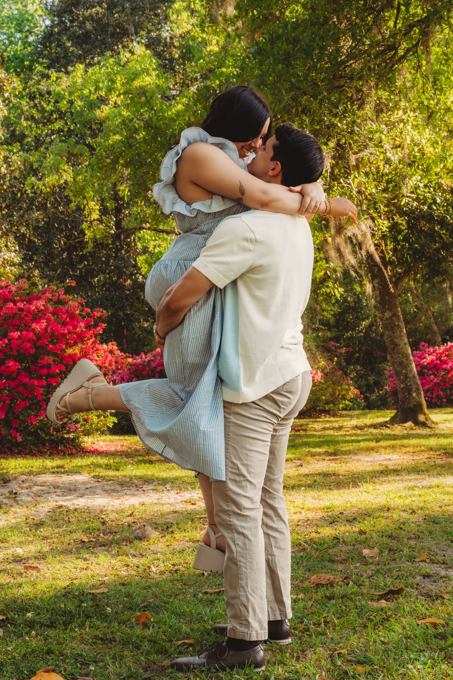 Couple sharing a romantic kiss in a lush garden, man lifting woman in a light blue dress among colorful flowers.