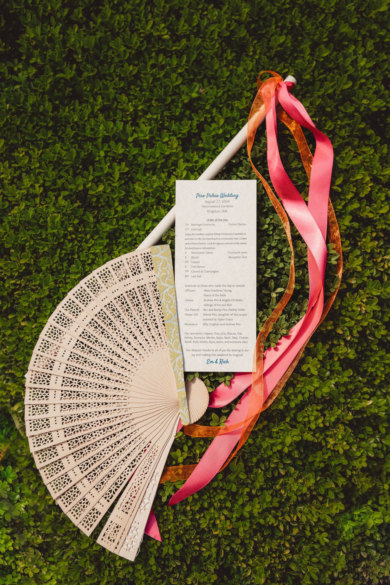Elegant wooden hand fan with lace cutwork beside a wedding program and pink ribbons on green grass.