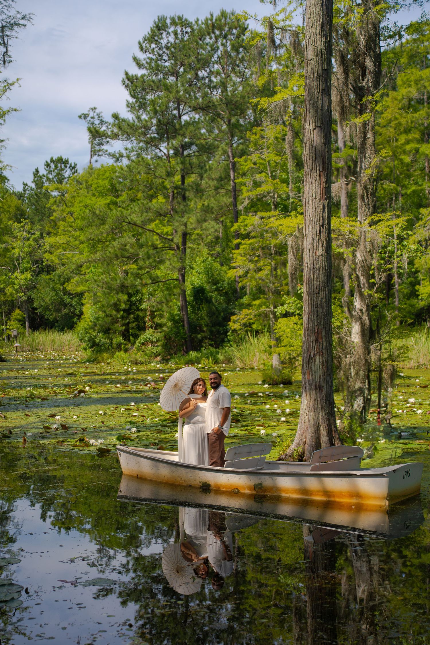 Couple in white standing in a boat on a serene lily pad lake surrounded by lush green cypress trees.
