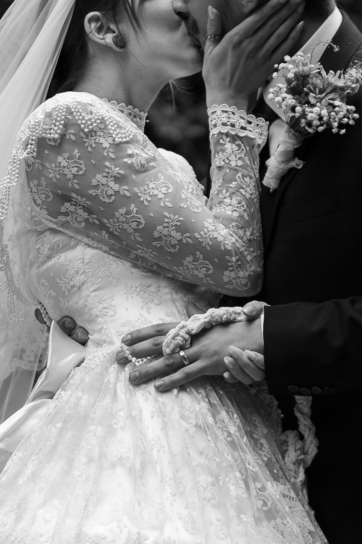 Black and white wedding photo of bride in lace gown and groom kissing, hands intertwined with wedding rings visible.