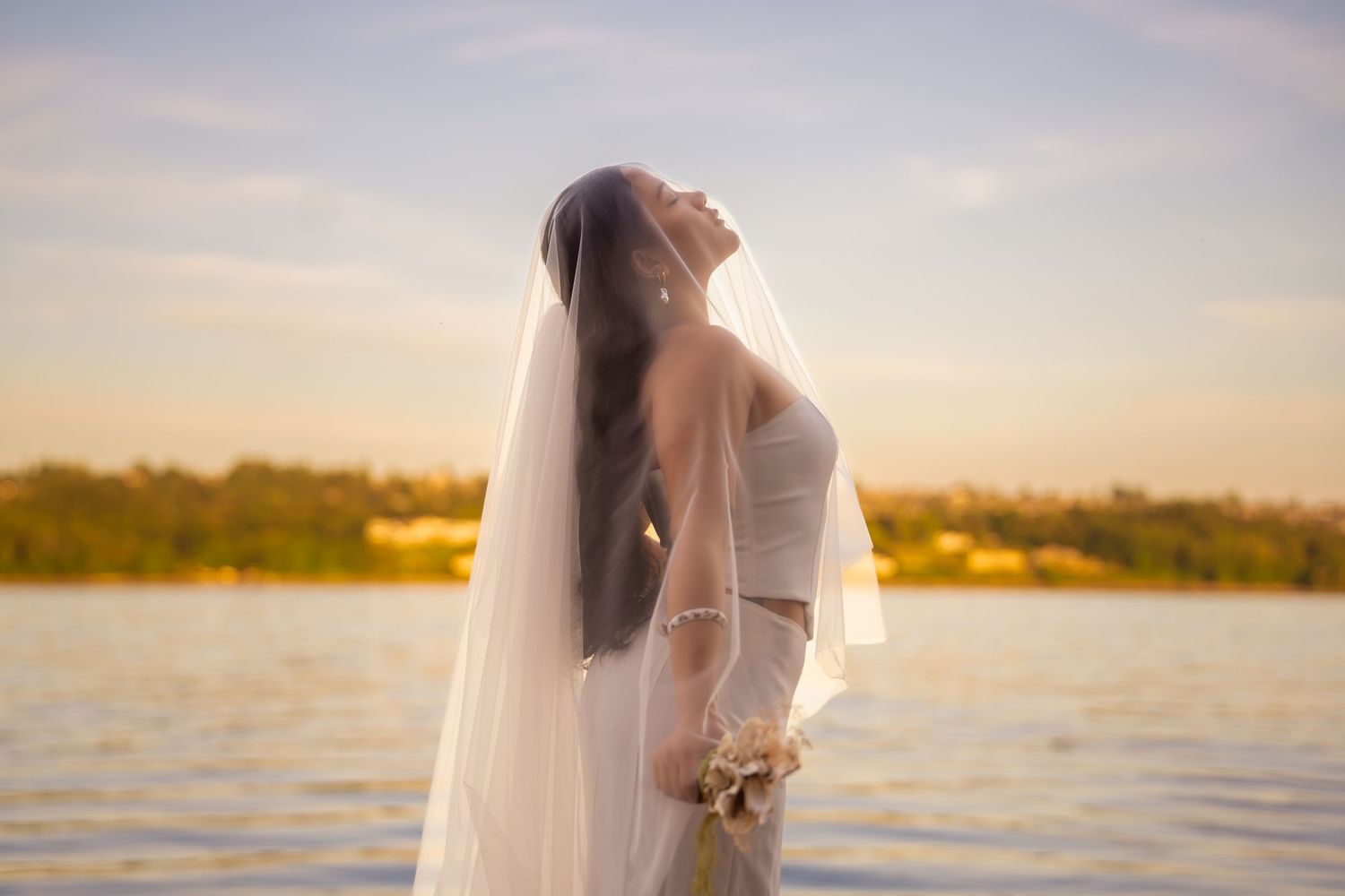 Bride in white gown and veil holding bouquet, head tilted back by waterfront at golden sunset.