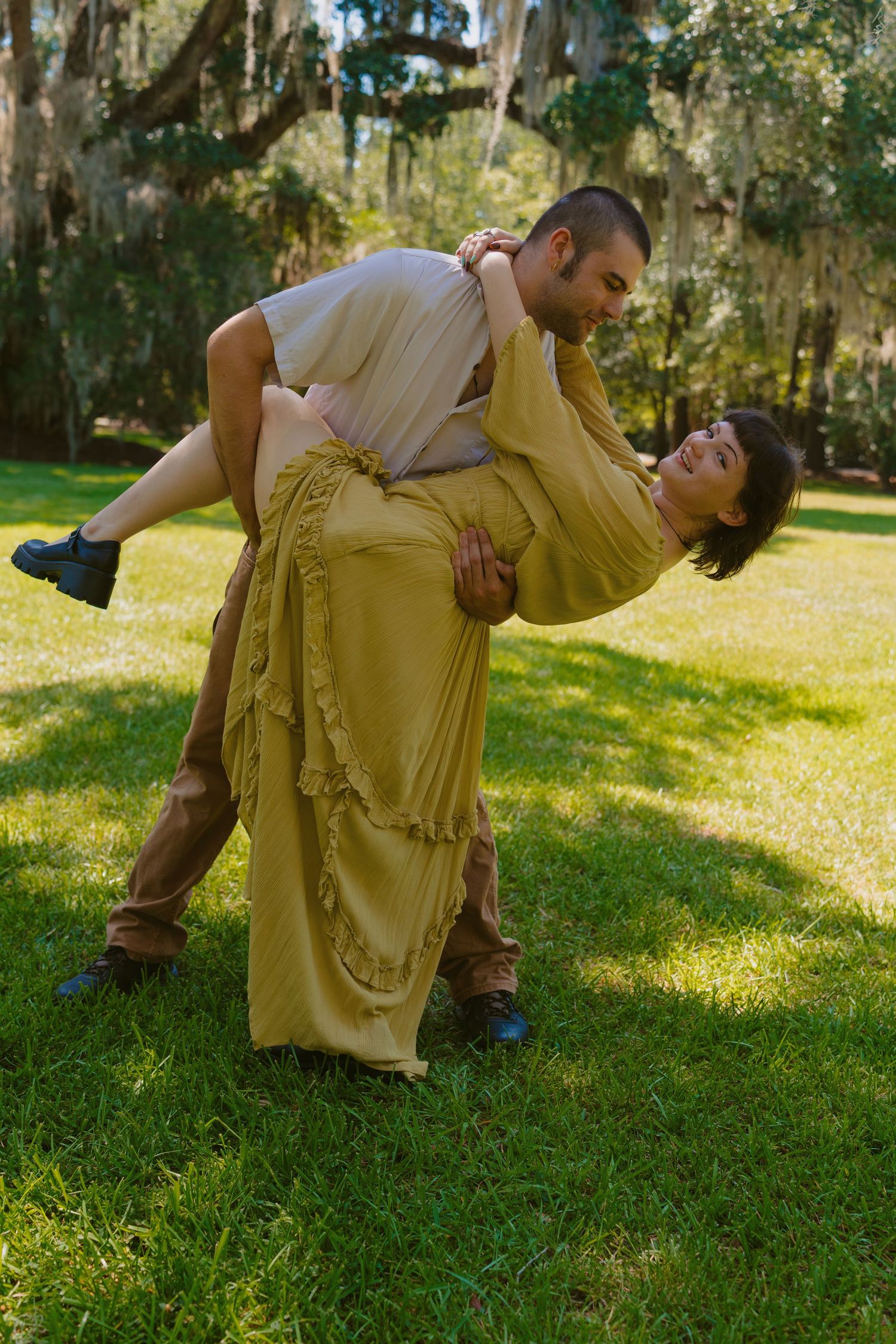 Couple dancing outdoors on green grass, man lifting woman in yellow dress near moss-covered trees.