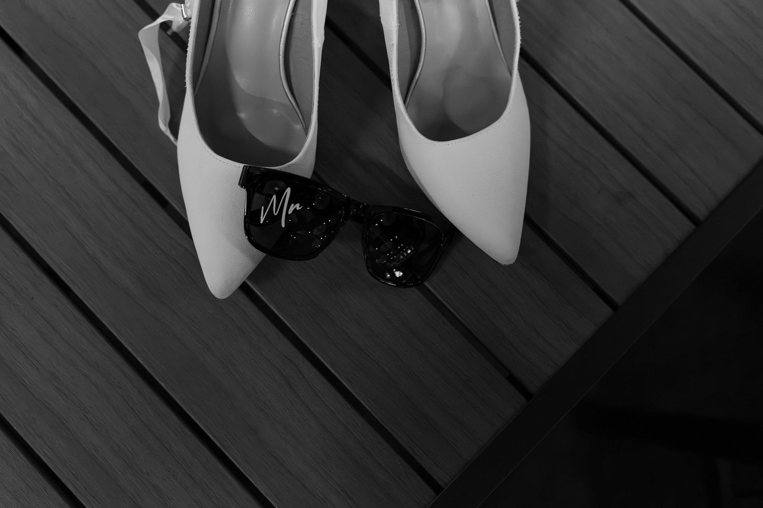 Black and white photo of pointed-toe bridal heels beside sunglasses with a Mr. charm, resting on a wooden deck.